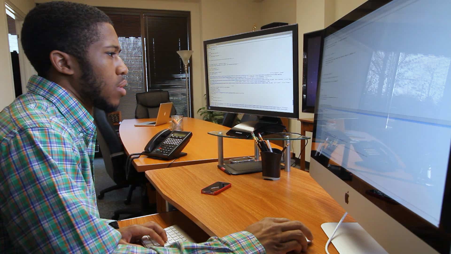 A person sits at a desk in an office, working on a computer with code displayed on two large monitors. The workspace has office supplies, a phone, and a laptop in the background.