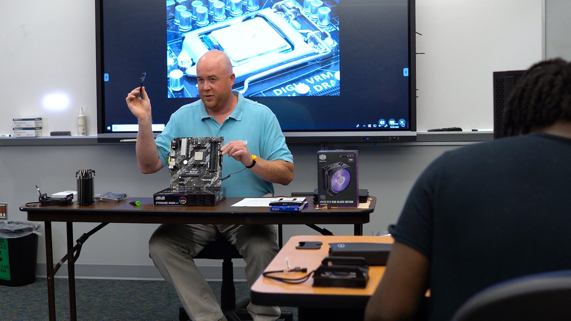 A teacher sits at a desk holding a computer component, with computer parts and tools laid out before him. A large screen behind displays a close-up of a motherboard. A student is seated in the foreground.