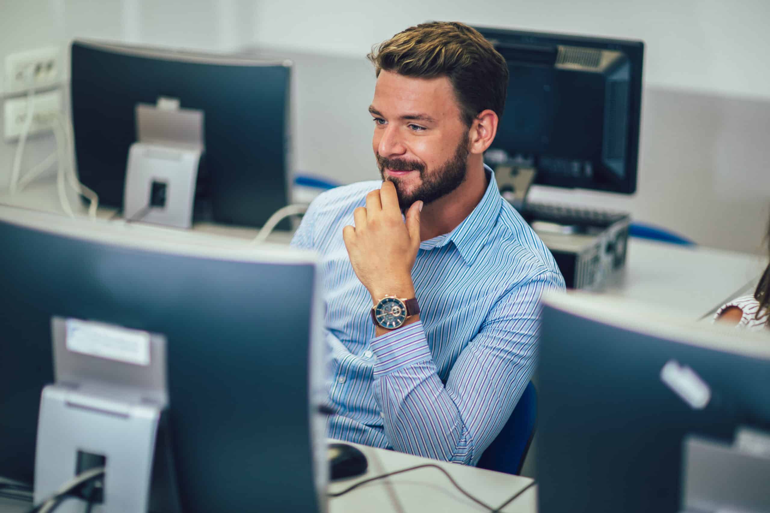 A man with a beard and striped shirt sits at a desk in an office, smiling and looking at a computer monitor, with other computer stations visible in the background.