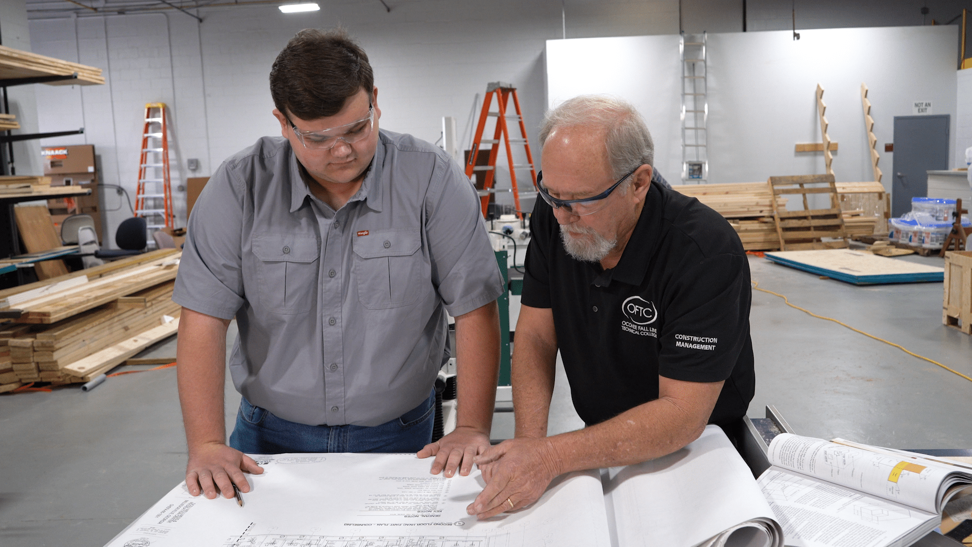 Two men wearing safety glasses look over architectural blueprints in a workshop. Shelves, ladders, and wooden materials are visible in the background, suggesting a construction or woodworking environment.
