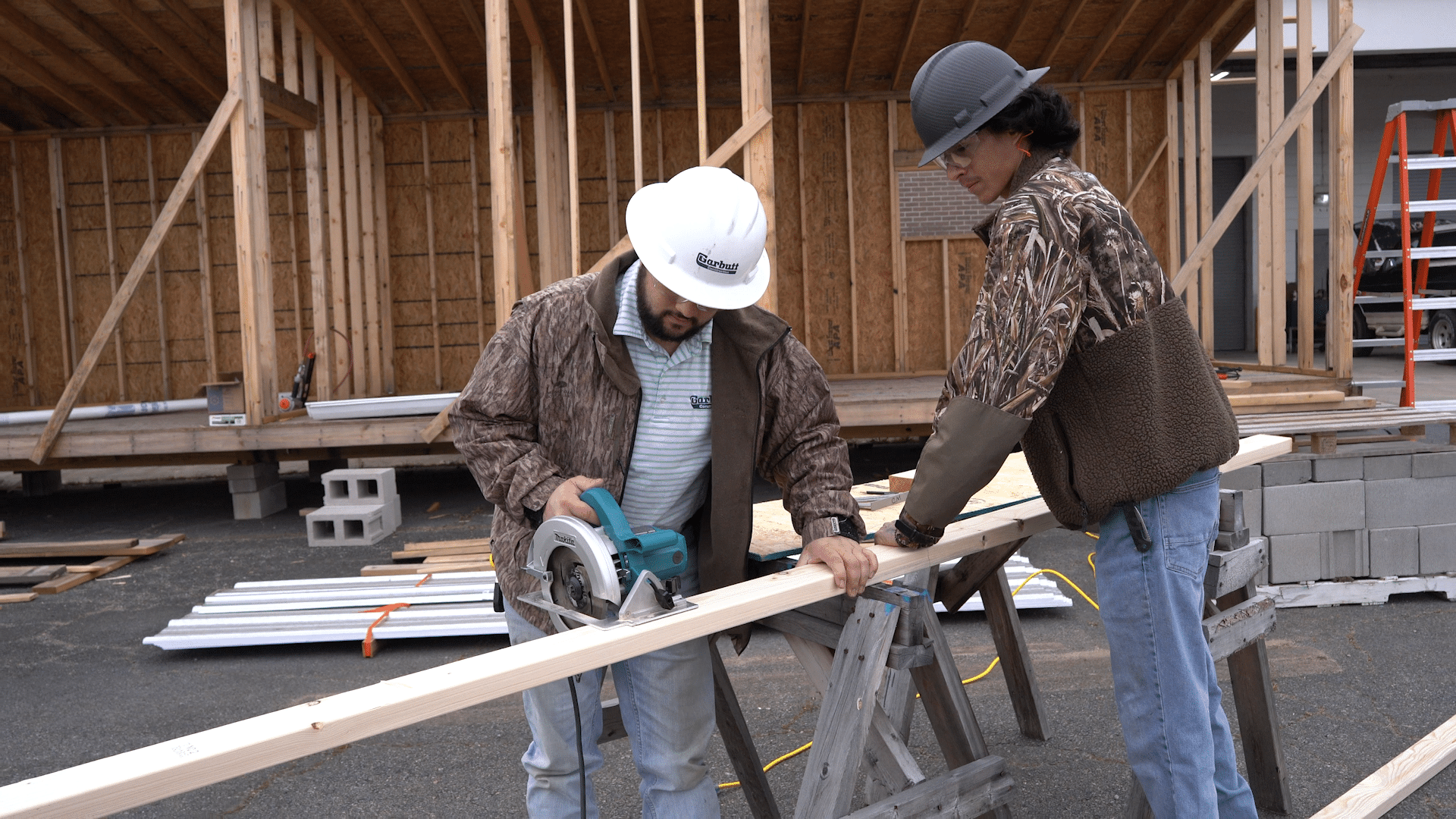 Two men wearing hard hats and safety gear use a circular saw to cut a wooden plank at a construction site with a partially built wooden structure and construction materials in the background.