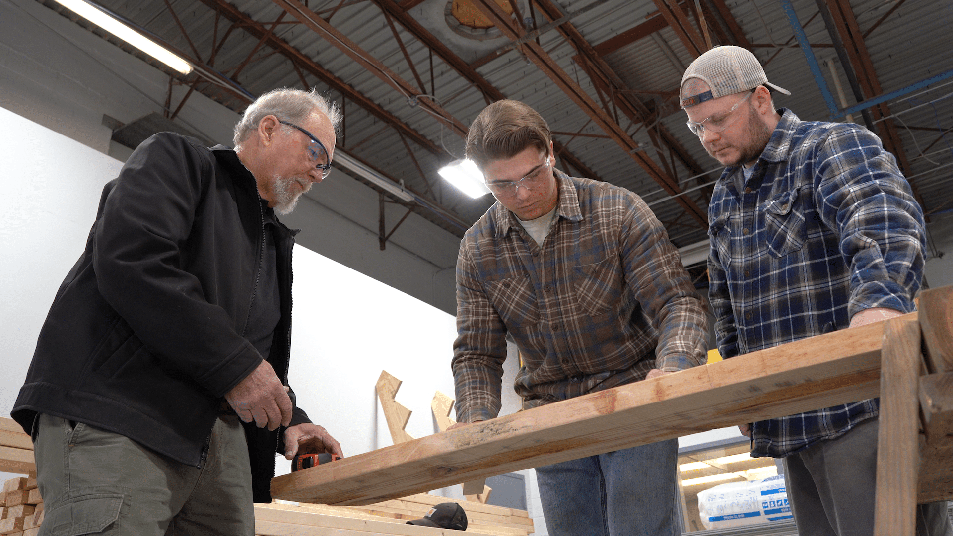 Three men wearing safety glasses and casual work clothes examine and measure a wooden beam in a workshop with a high ceiling and exposed pipes.