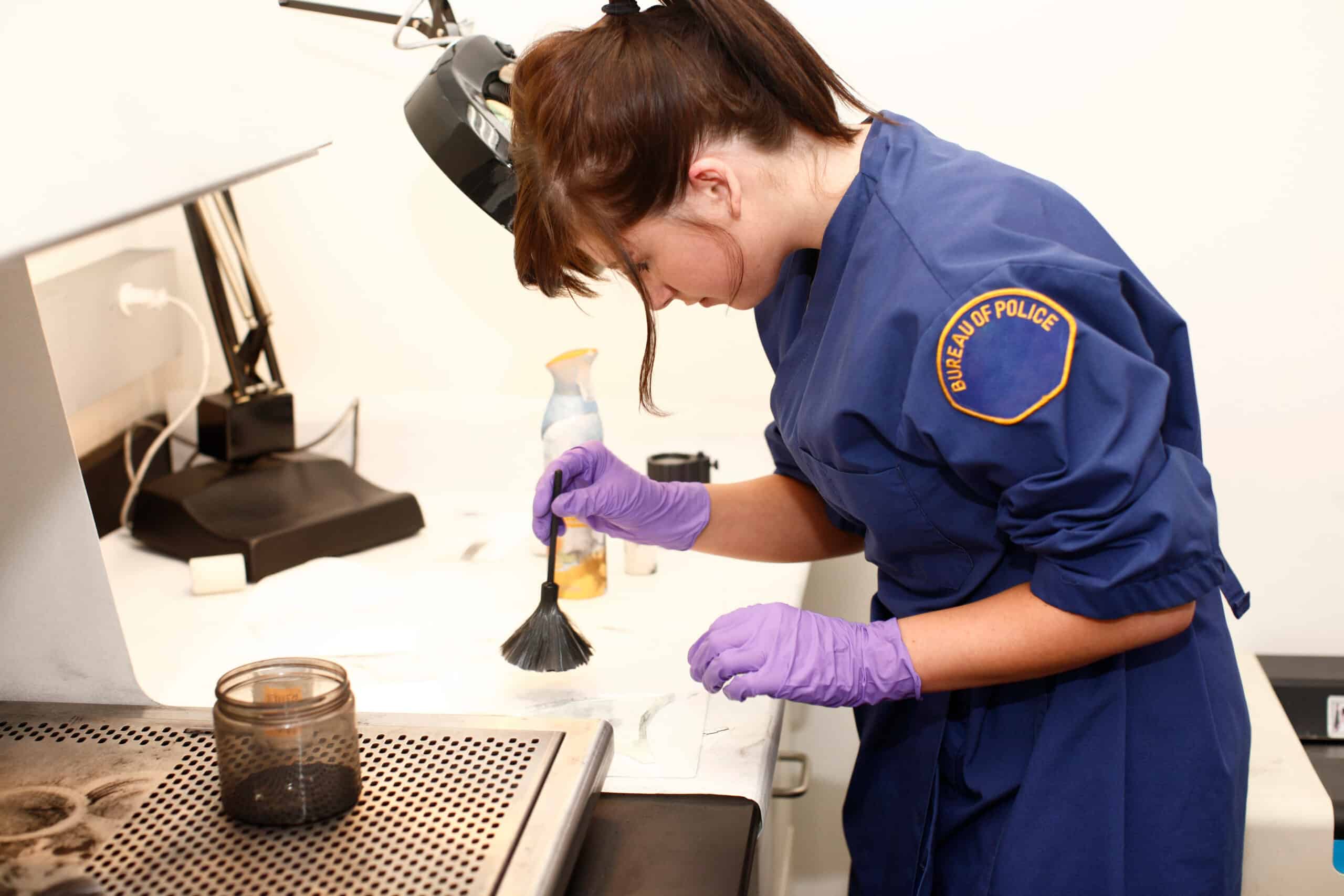 A police officer wearing a blue uniform and purple gloves uses a brush to dust for fingerprints on a surface in a forensic lab.