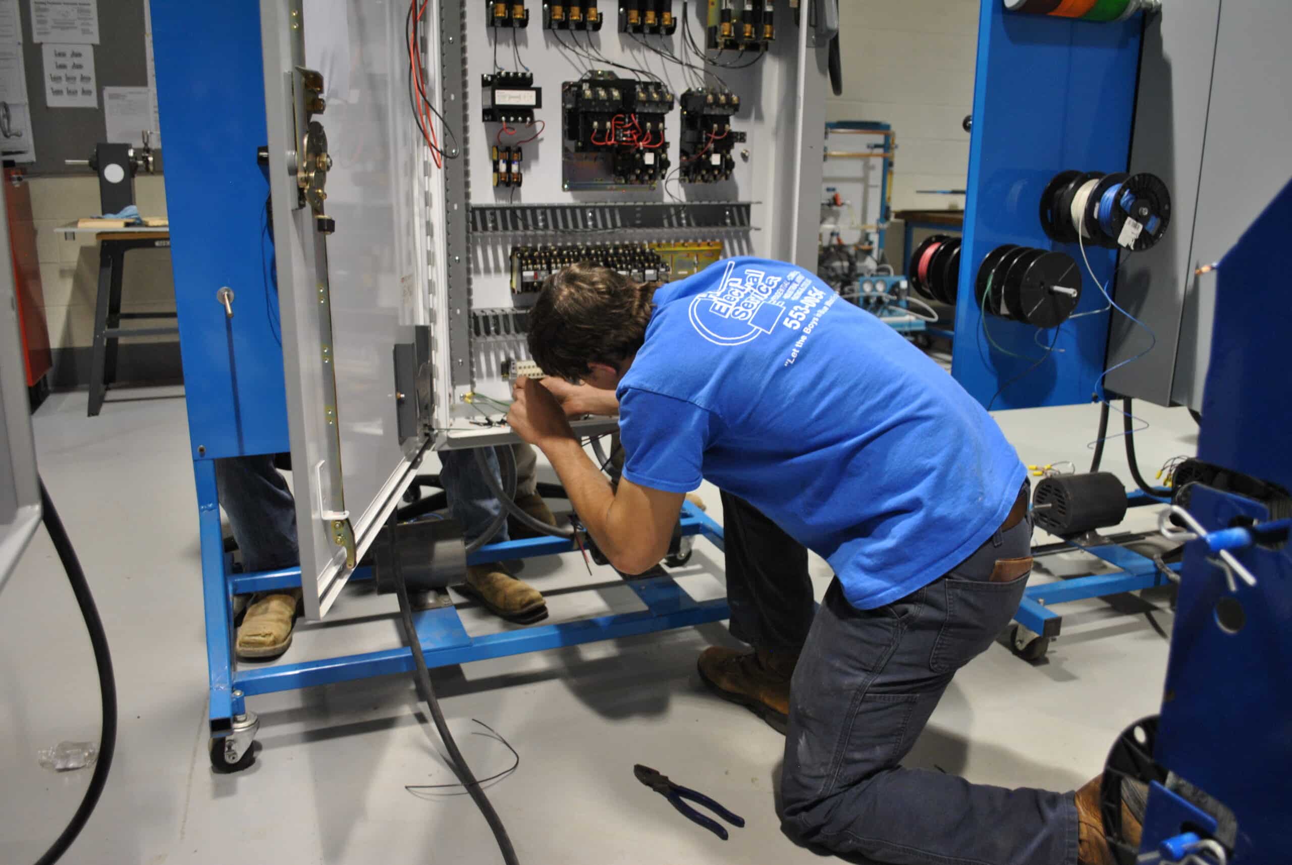 A person in a blue shirt kneels while working on wiring inside an open electrical control panel, surrounded by tools and cables in an industrial workshop setting.