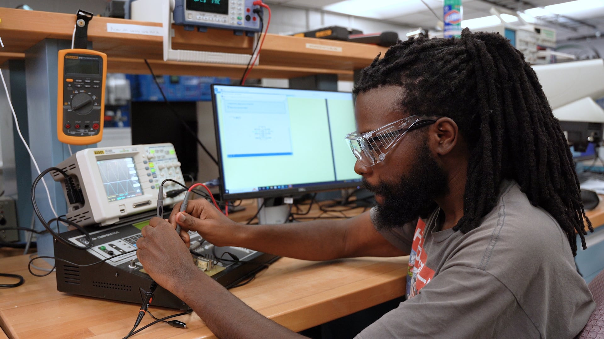 A person wearing safety glasses works at a lab bench with electronic equipment using probes on a device while computer monitors and measuring instruments display data in the background.