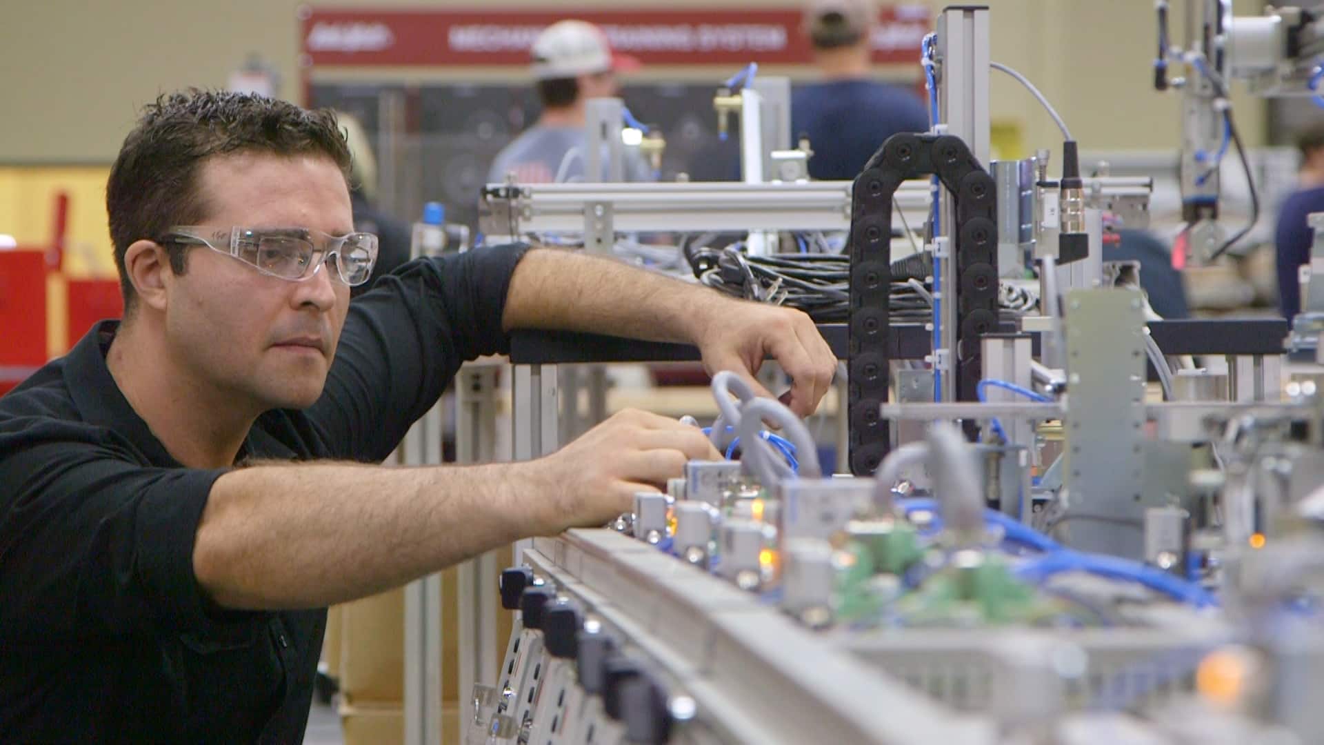 A man wearing safety glasses adjusts components on an industrial machine in a workshop, focusing intently while surrounded by cables, circuit boards, and other equipment.
