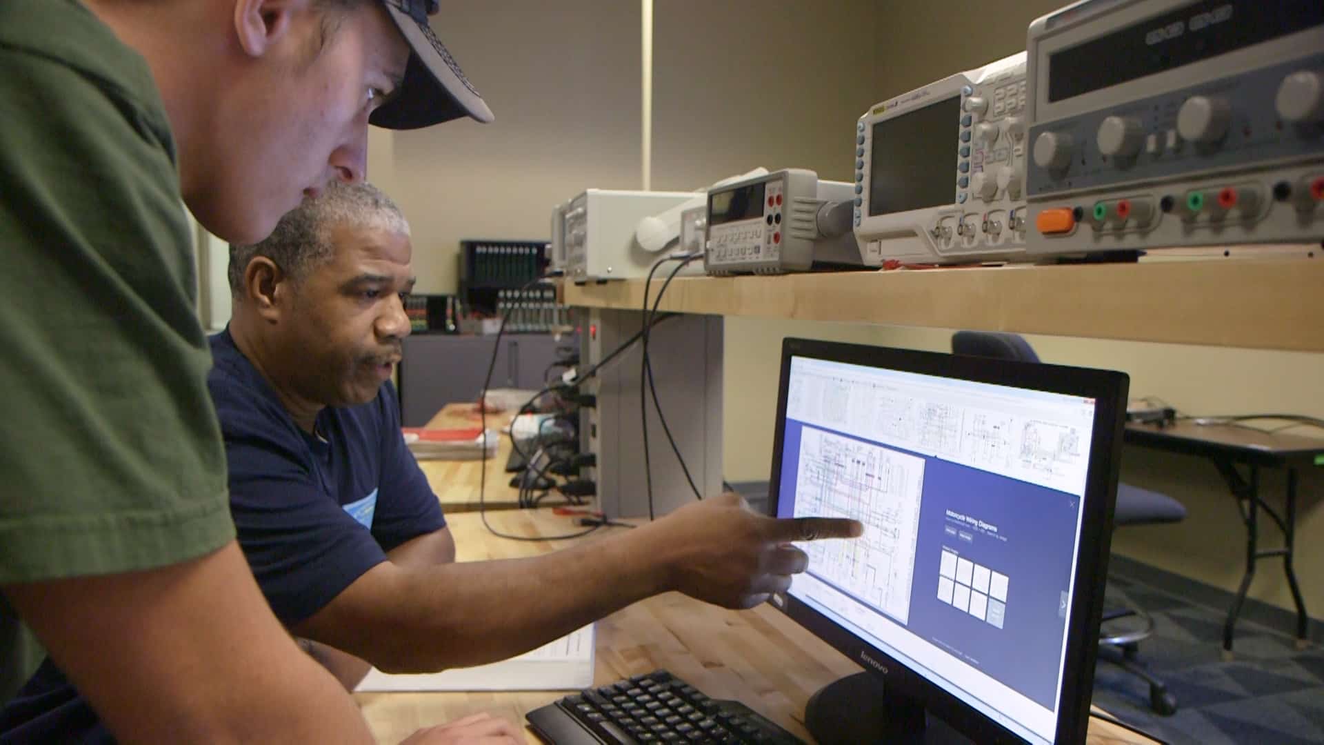 Two men work together at a desk with electronics equipment, looking at technical diagrams on a computer monitor. One man is seated while the other stands beside him, pointing at the screen.