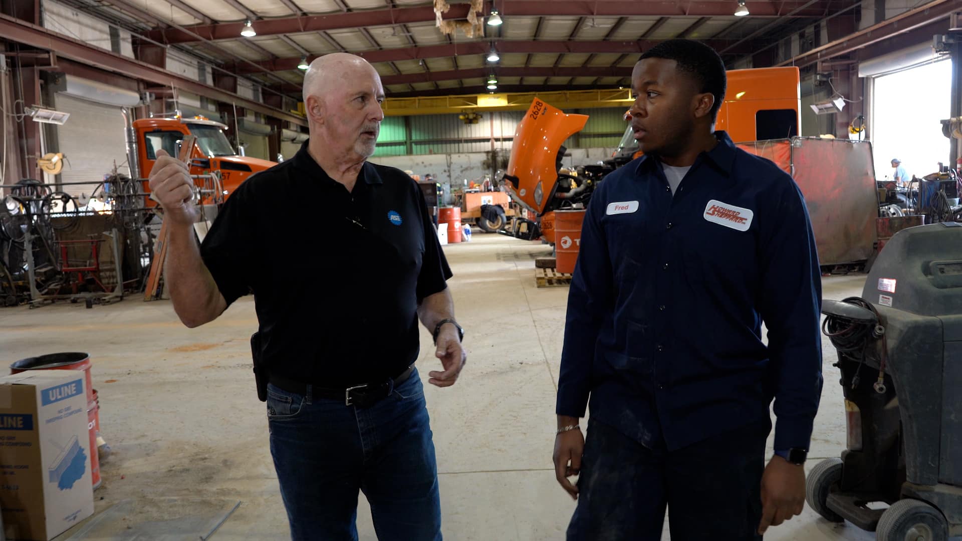 Two men stand talking in a large industrial workshop with various tools, equipment, and orange trucks in the background. One man gestures while speaking, and the other listens attentively.
