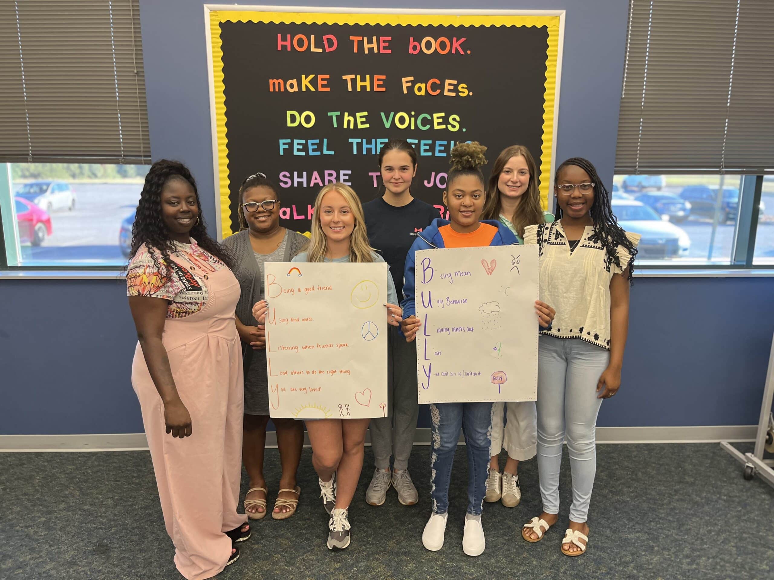 A group of seven smiling students stand indoors, holding colorful anti-bullying posters. A bulletin board behind them reads: HOLD THE BOOK. MAKE THE FACES. DO THE VOICES. FEEL THE CHARACTER. SHARE THE STORY.