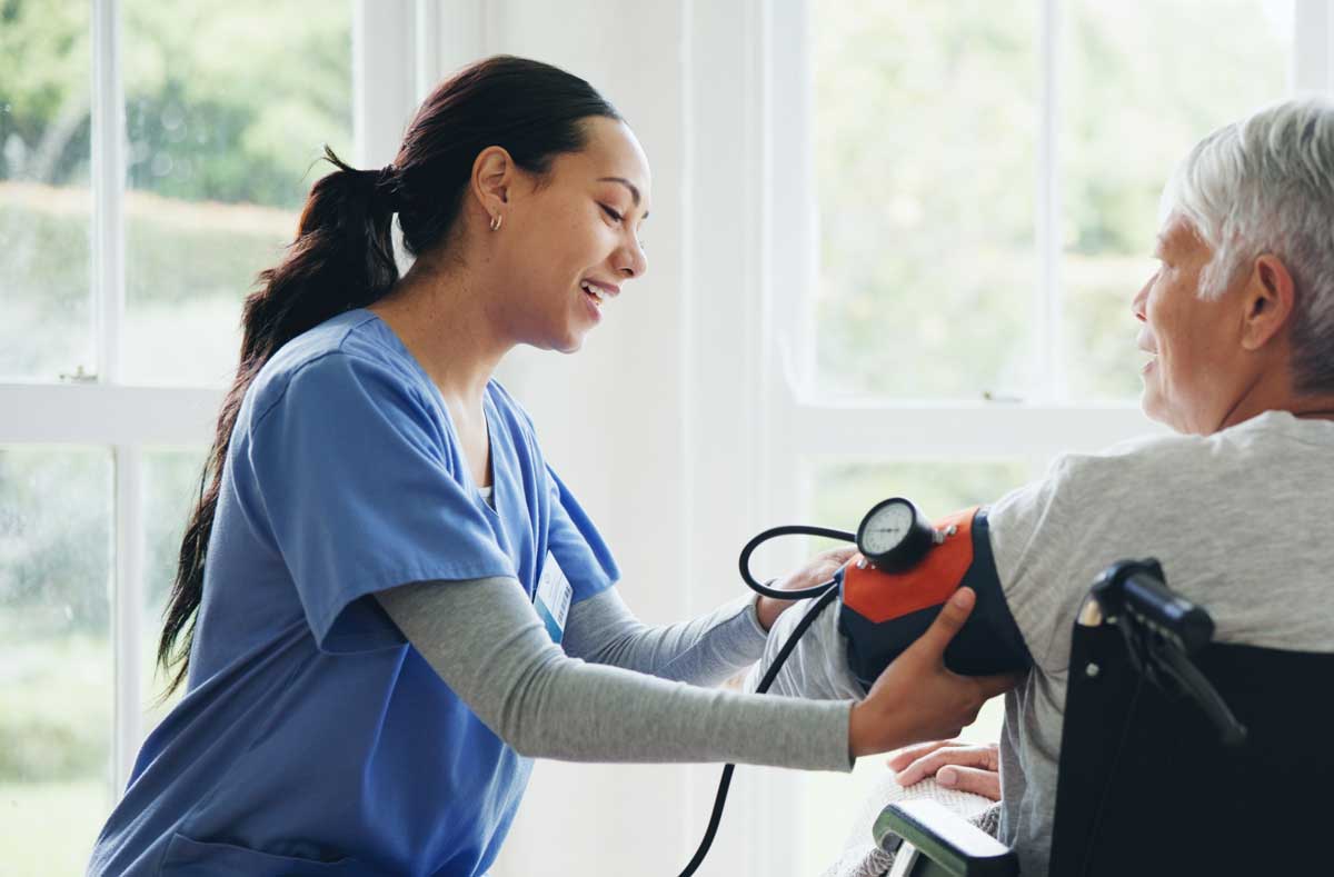 A nurse in blue scrubs smiles while measuring the blood pressure of an older adult seated in a wheelchair near a bright window.