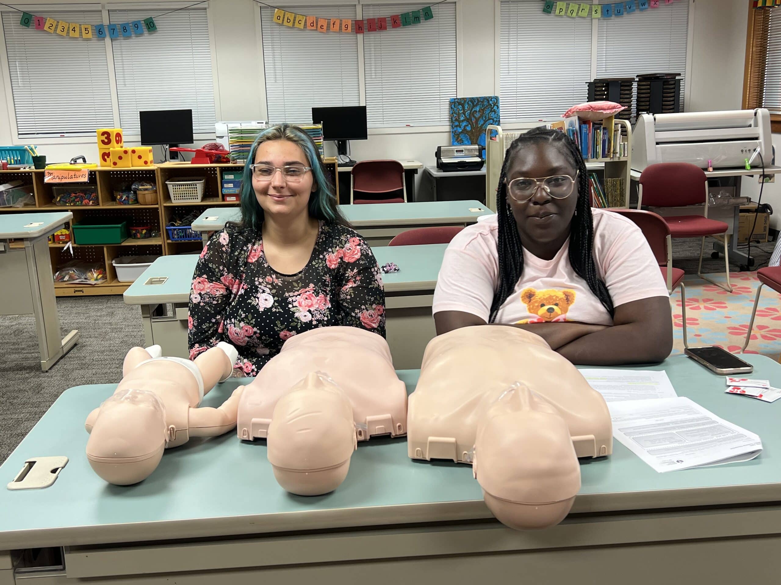 Two women sit at a table in a classroom with three CPR training mannequins an infant and two adults in front of them. Papers and desks are visible in the background.