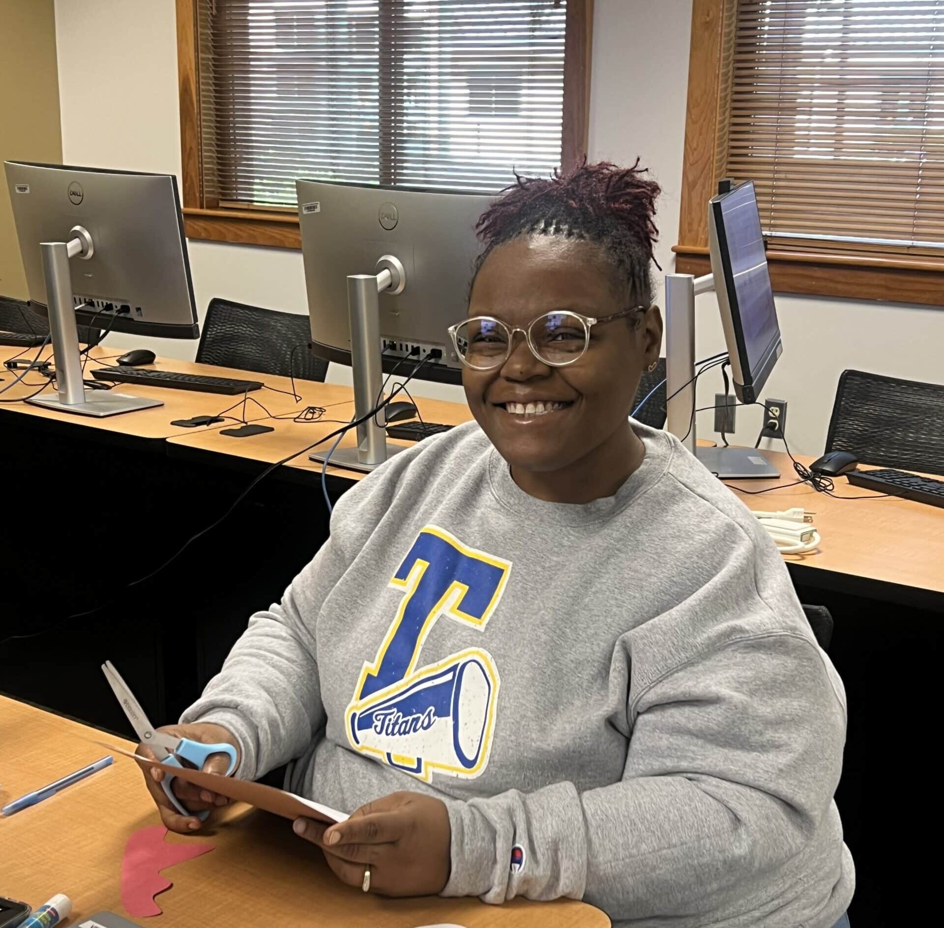 A person wearing glasses and a gray Titans sweatshirt smiles while using scissors at a desk in a computer lab. Laptops, papers, and desktop computers are visible in the background.