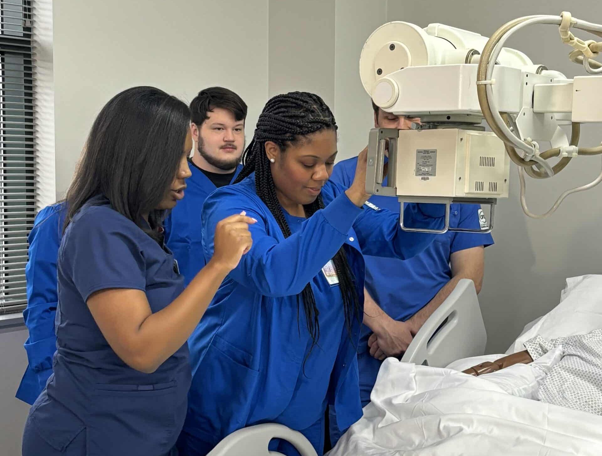 A group of medical professionals or students in blue scrubs stand around a hospital bed, positioning an X-ray machine over a patient covered with a white sheet.