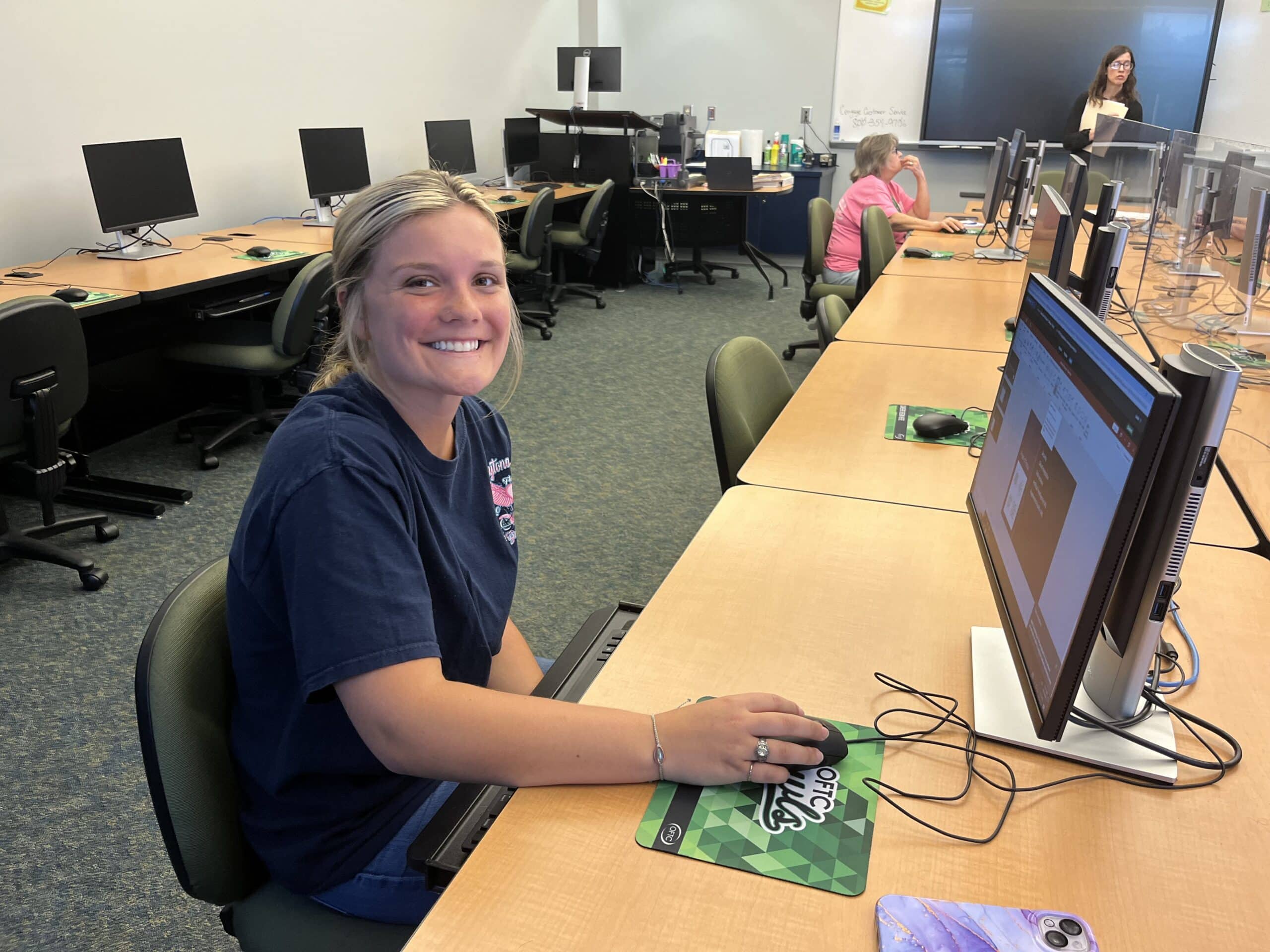 A young woman smiles at the camera while seated at a computer in a classroom. Other students and a teacher are visible in the background, with computers on desks around the room.