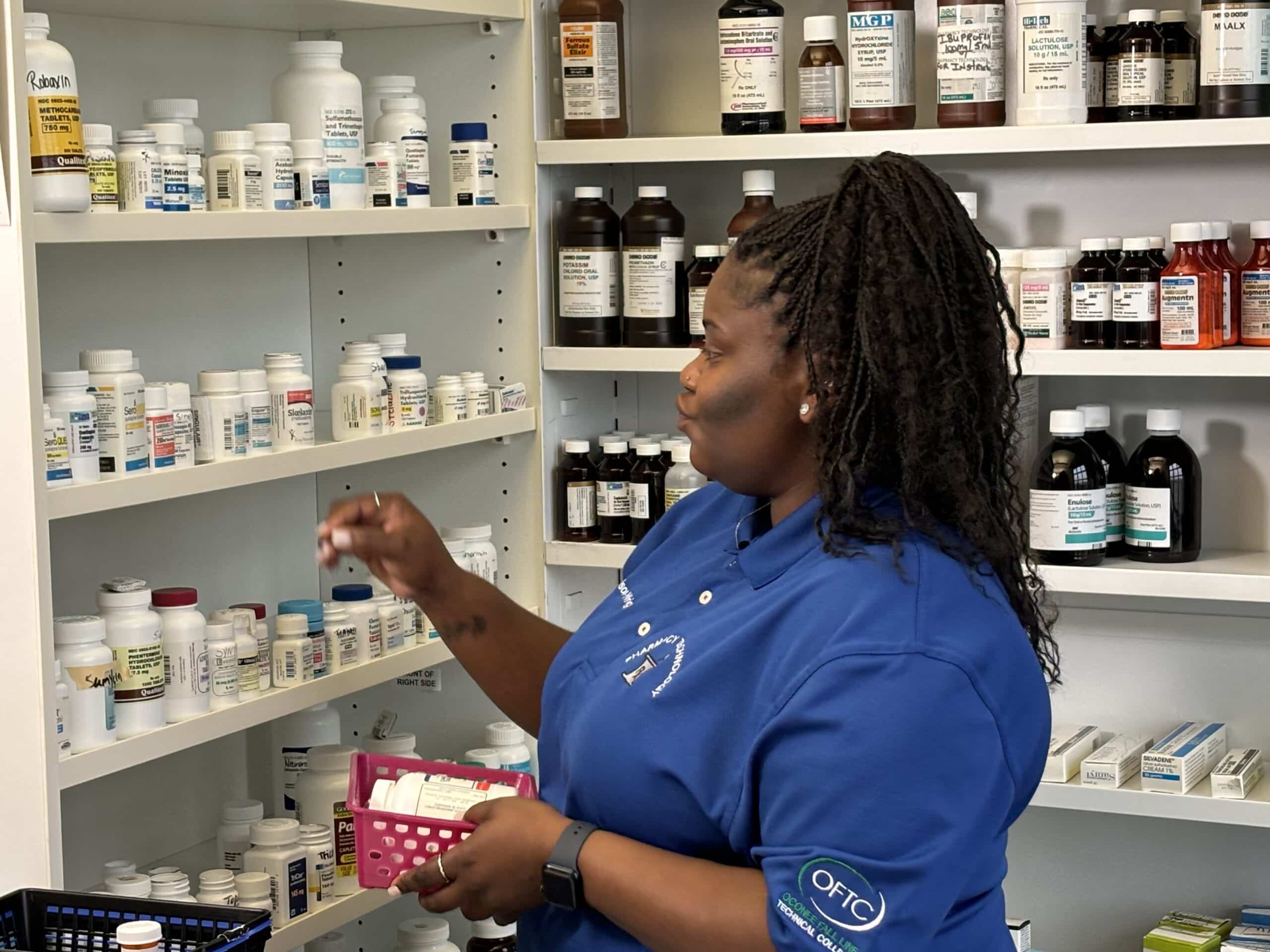 A woman in a blue shirt organizes medication bottles on shelves in a pharmacy, holding a small pink basket filled with prescription containers.