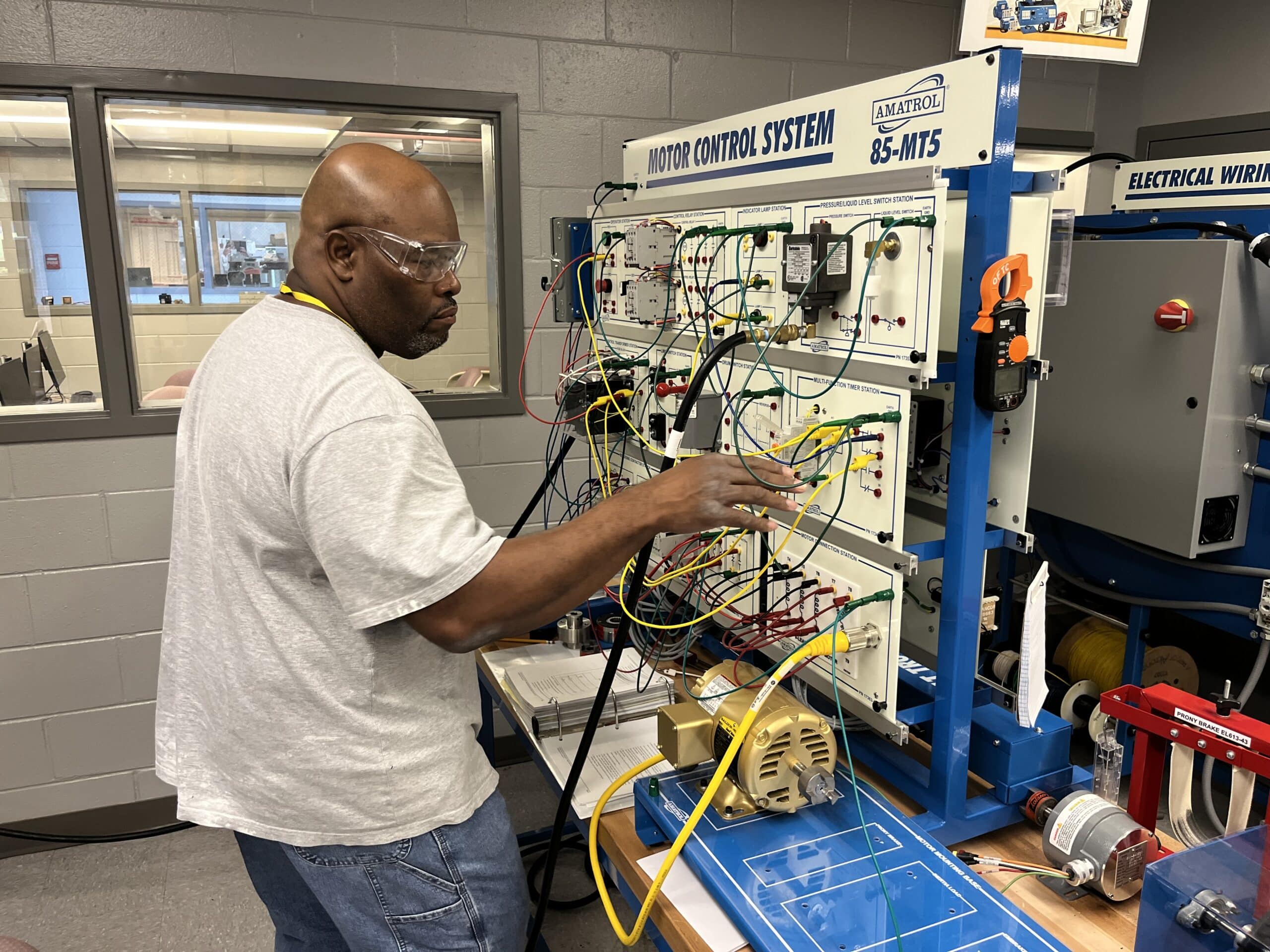 A man wearing safety glasses works with a motor control system training panel, adjusting wires and components in a technical lab setting with gray walls and observation windows.