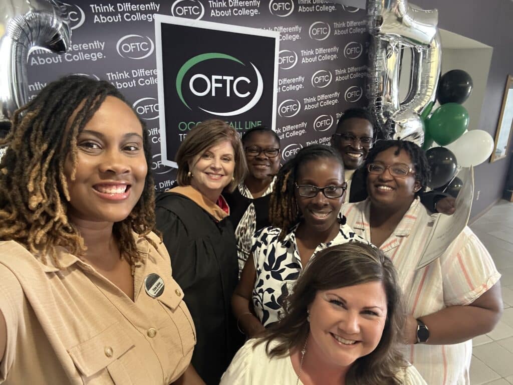 A group of seven smiling adults pose indoors at an OFTC event, standing in front of a branded backdrop and large silver 5 balloon. Some wear business attire, and one person is in academic regalia.