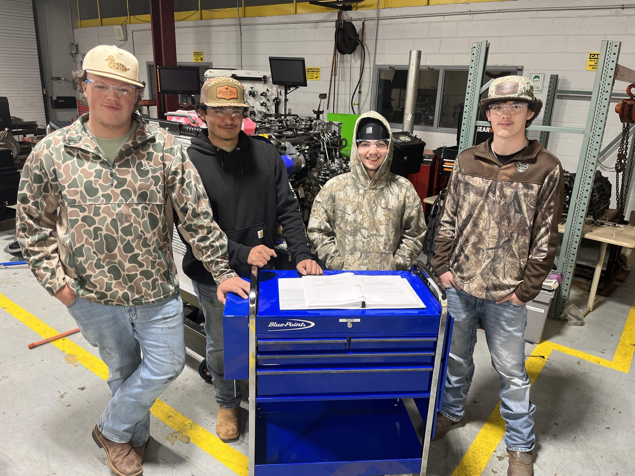 Four young men wearing safety glasses, hats, and camouflage or work clothes stand around a blue tool chest in an industrial workshop, smiling at the camera. Papers are spread out on the tool chest.