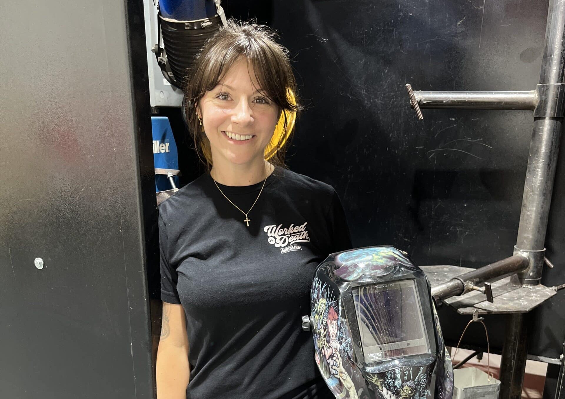 A woman wearing a black Wicked Welders t-shirt and gray pants stands in a workshop, smiling at the camera. She is holding a decorated welding helmet. Industrial equipment is visible in the background.