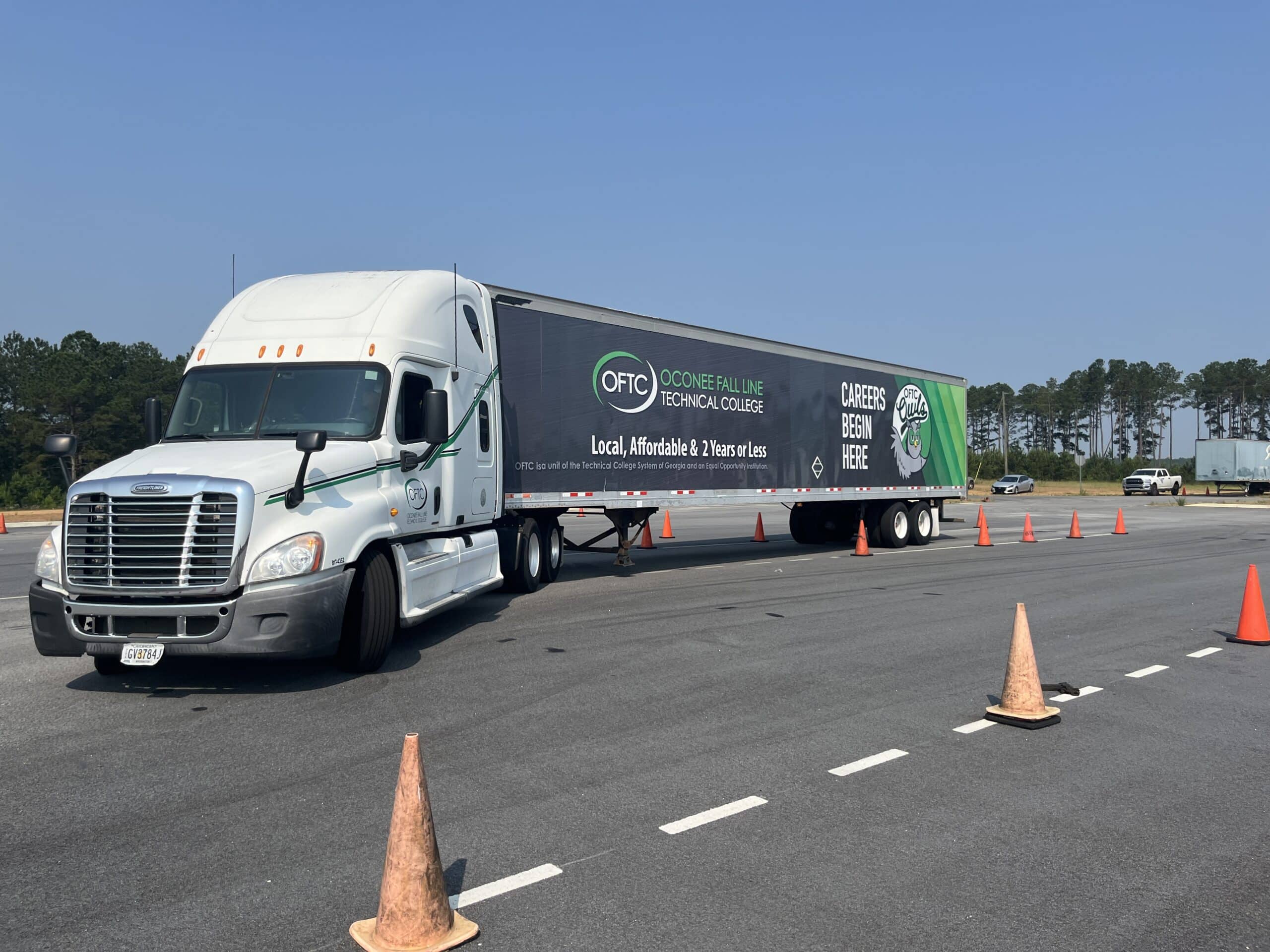 A white semi-truck with an Oconee Fall Line Technical College trailer is parked on an asphalt lot, surrounded by orange traffic cones, with trees and a blue sky in the background.