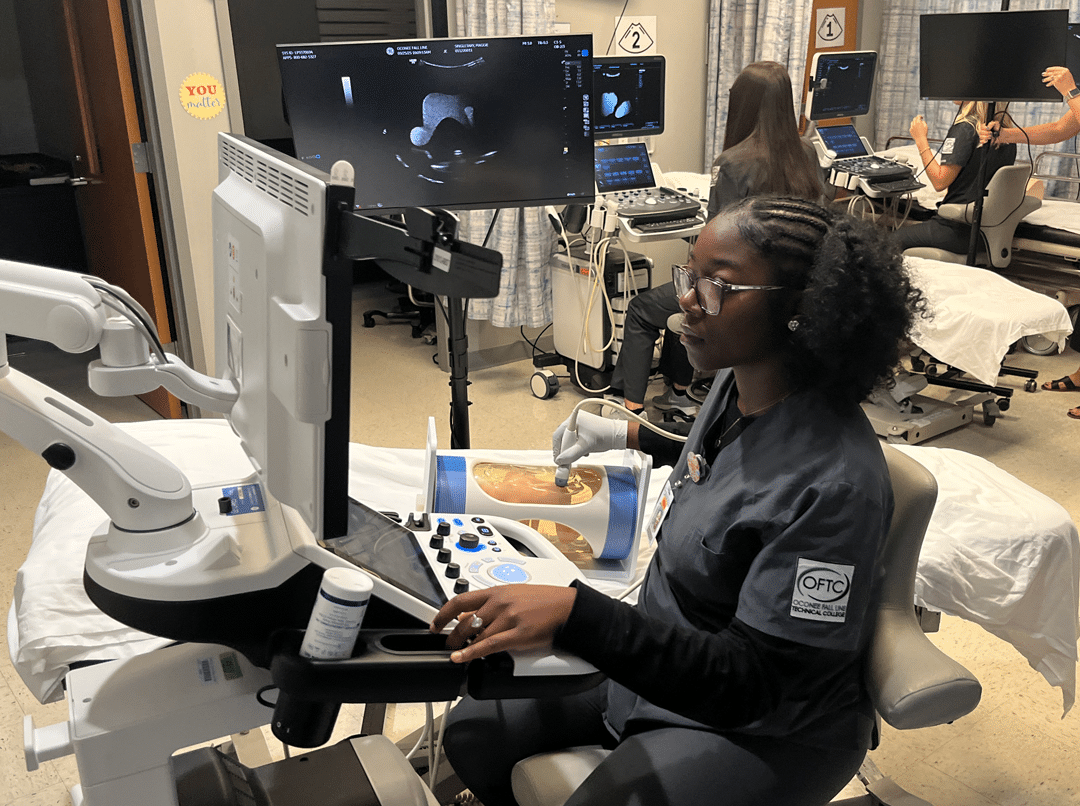 Student in scrubs using an ultrasound machine on a practice dummy as others train with similar devices in a clinical training room.