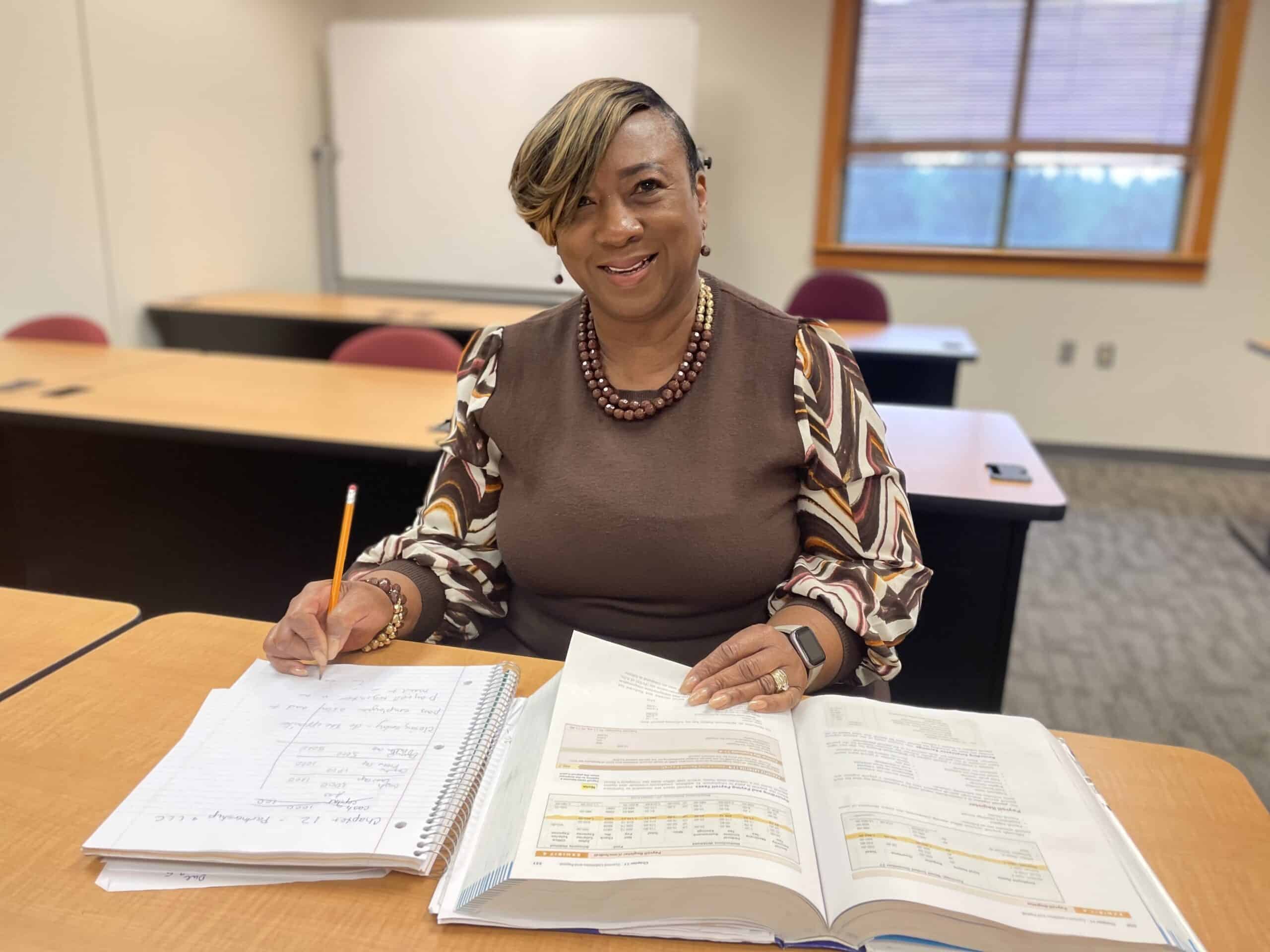 A woman sits at a classroom desk, smiling, with an open textbook and notebook in front of her. She holds a pencil and wears a brown top with patterned sleeves and a beaded necklace. Empty desks and a window are in the background.