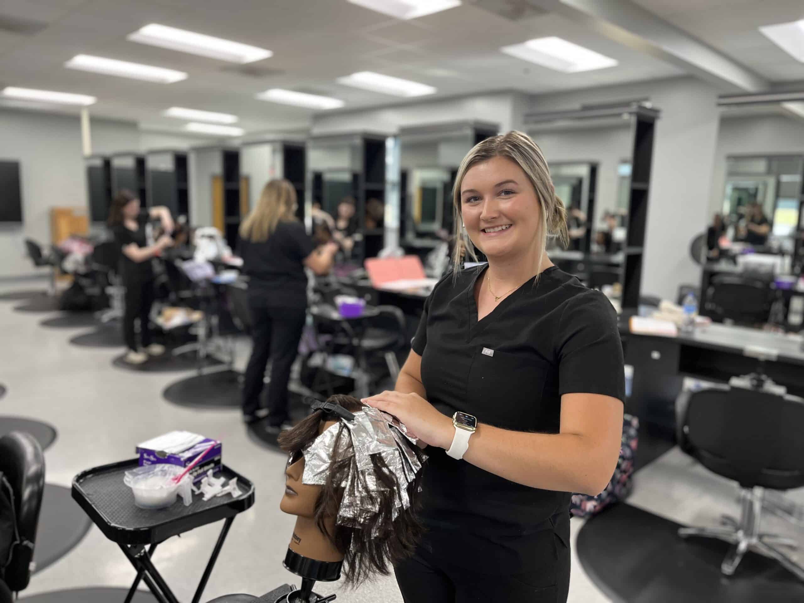 A smiling woman in black scrubs styles a mannequin head with foils on its hair in a modern salon classroom. Other people work in the background at salon stations.