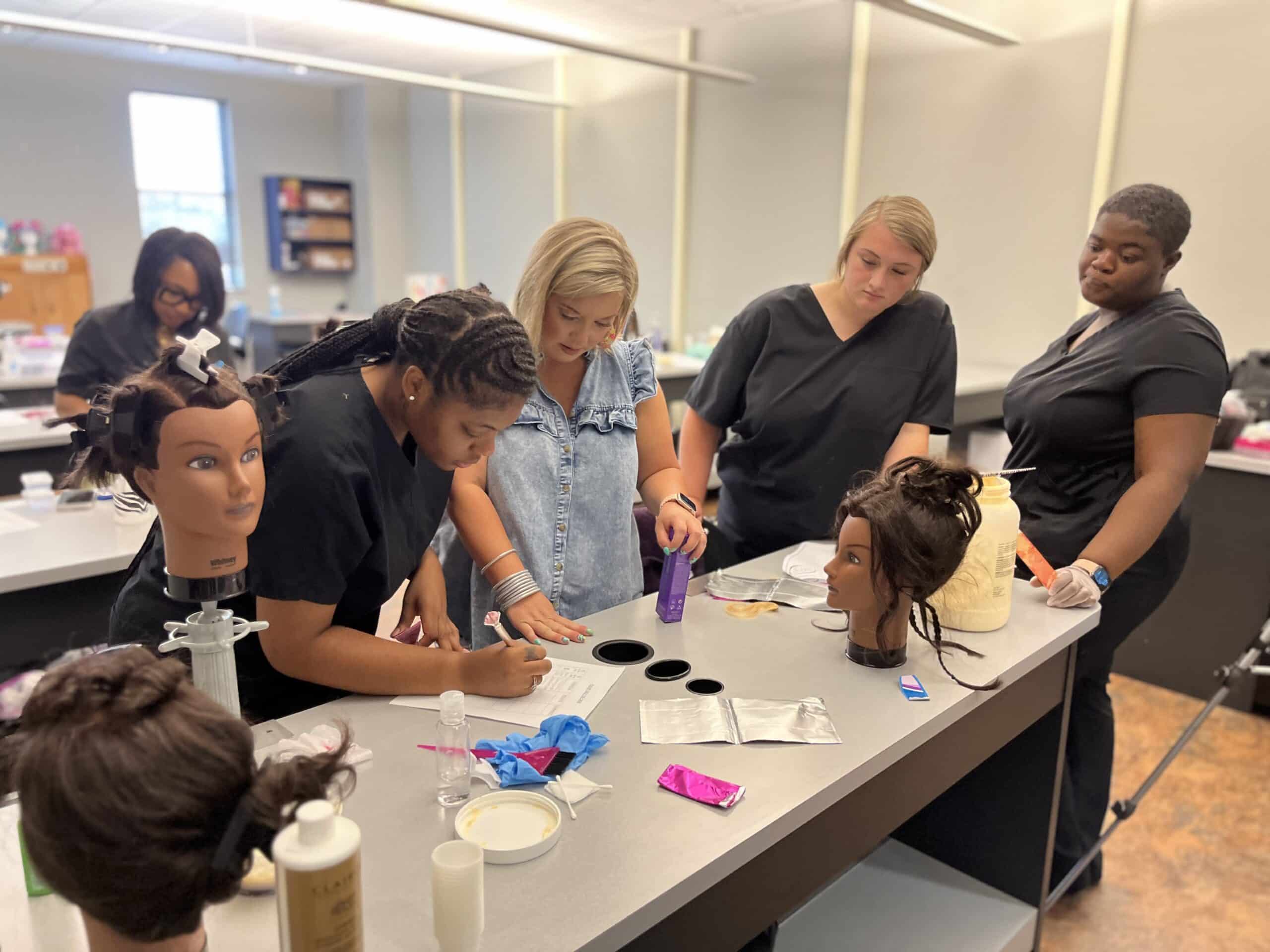 Five people in a classroom work around a table with hairstyling mannequins, hair products, and tools. One person writes on paper while others observe and discuss, practicing hair techniques in a cosmetology class.