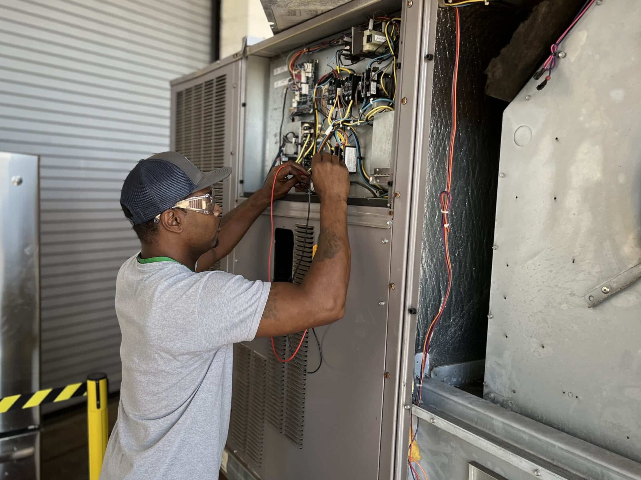 A man wearing safety glasses and a cap works on exposed wiring inside a large industrial electrical panel, with tools and wires visible around him. A yellow caution barrier is nearby.