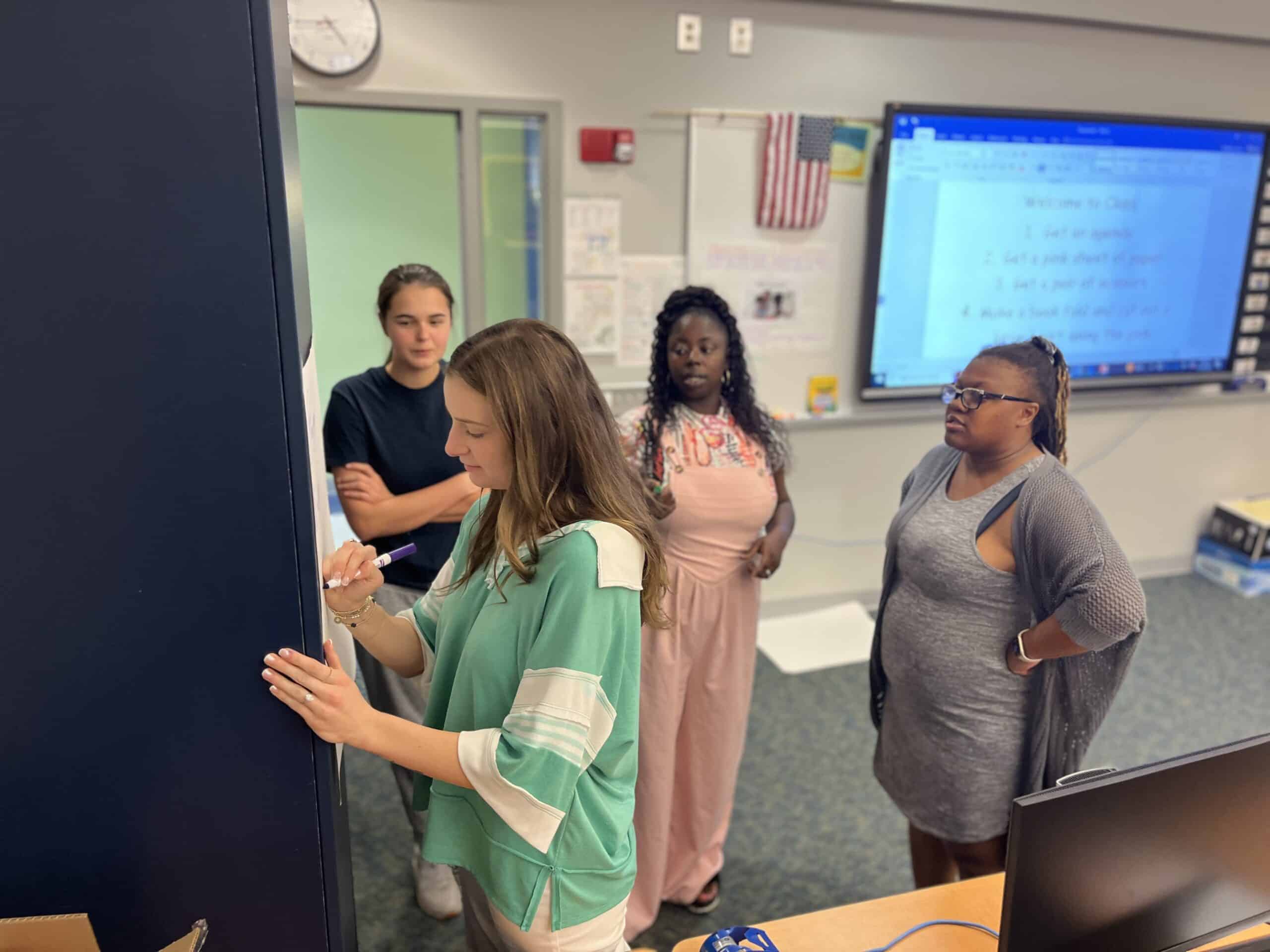 Four women stand in a classroom, with one writing on a board while the others observe. A screen with notes, an American flag, and a clock are visible in the background.