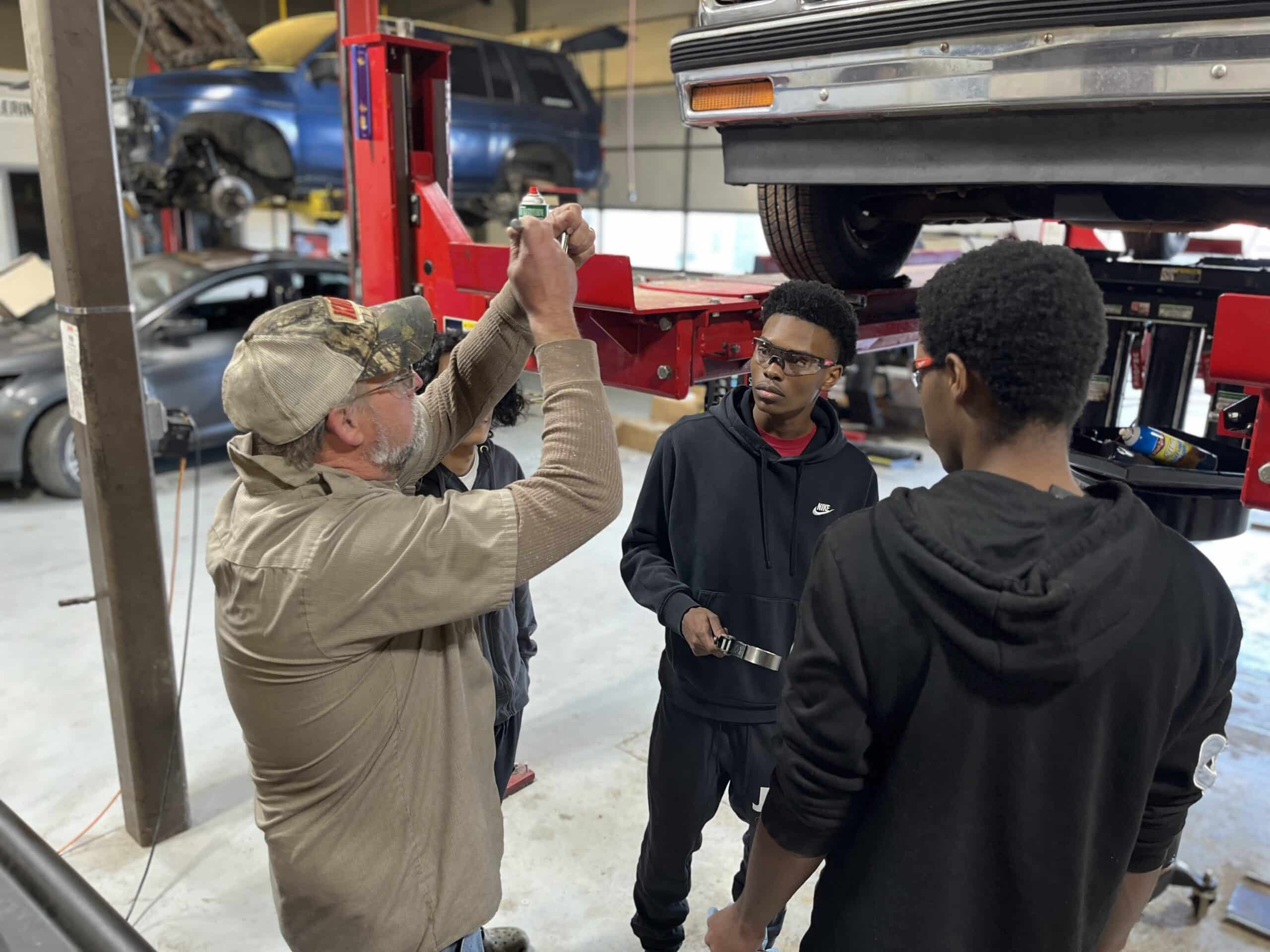 A mechanic explains a car part to three students in a garage. The group stands near a lifted vehicle, surrounded by tools and other cars in the background.