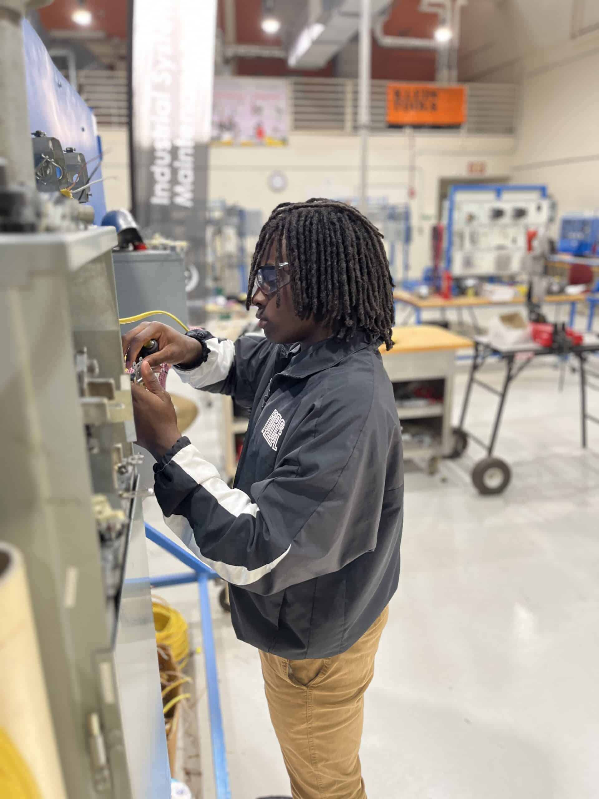 A person wearing safety glasses and a black jacket works with electrical equipment in a bright, industrial classroom filled with tools, machinery, and workstations.