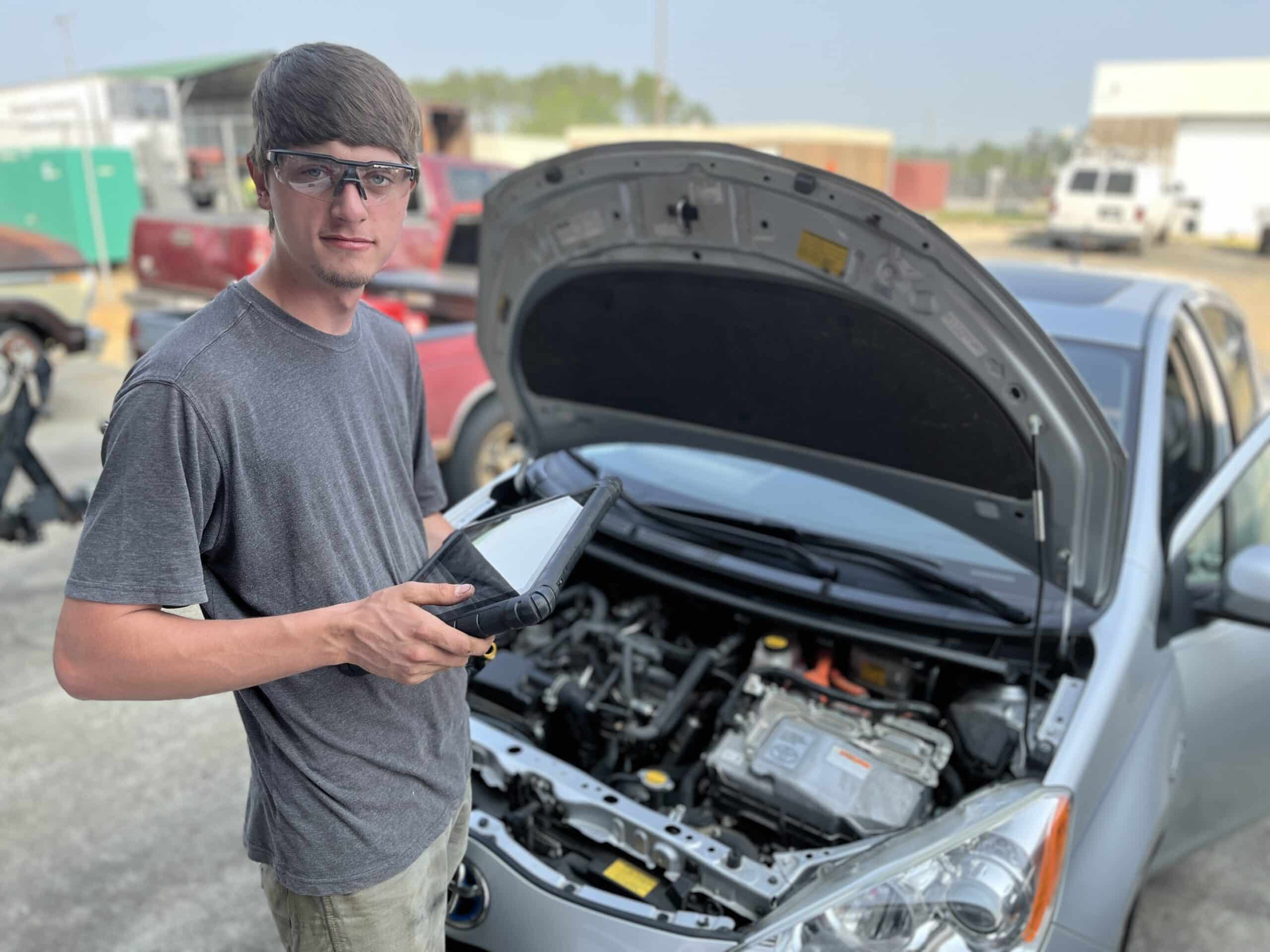 A young man wearing safety glasses holds a diagnostic tablet while standing next to a car with its hood open, inspecting the engine in an outdoor area with other vehicles in the background.