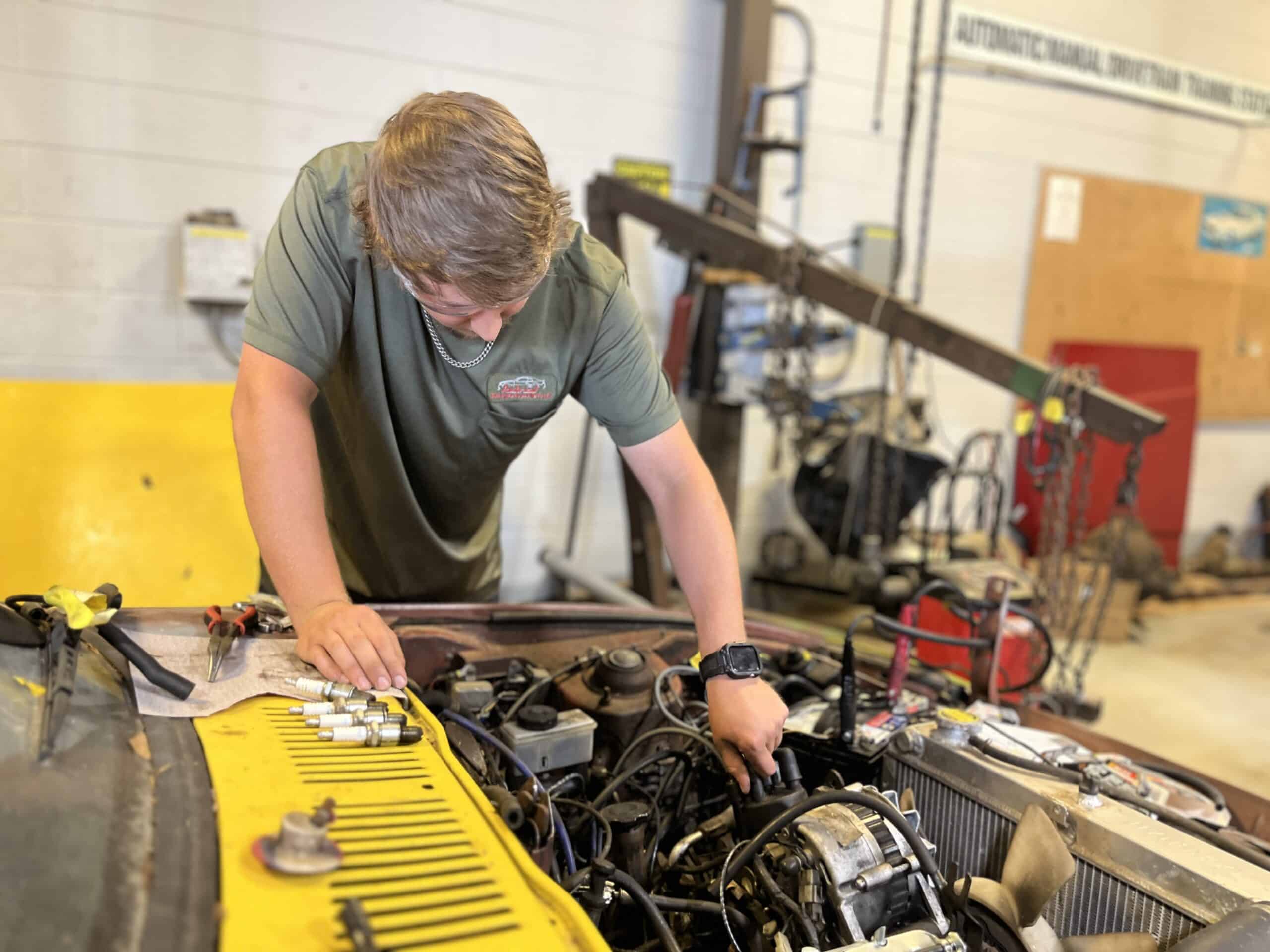A young man in a green shirt works on a car engine in a brightly lit auto repair shop, with various tools and engine parts scattered nearby.