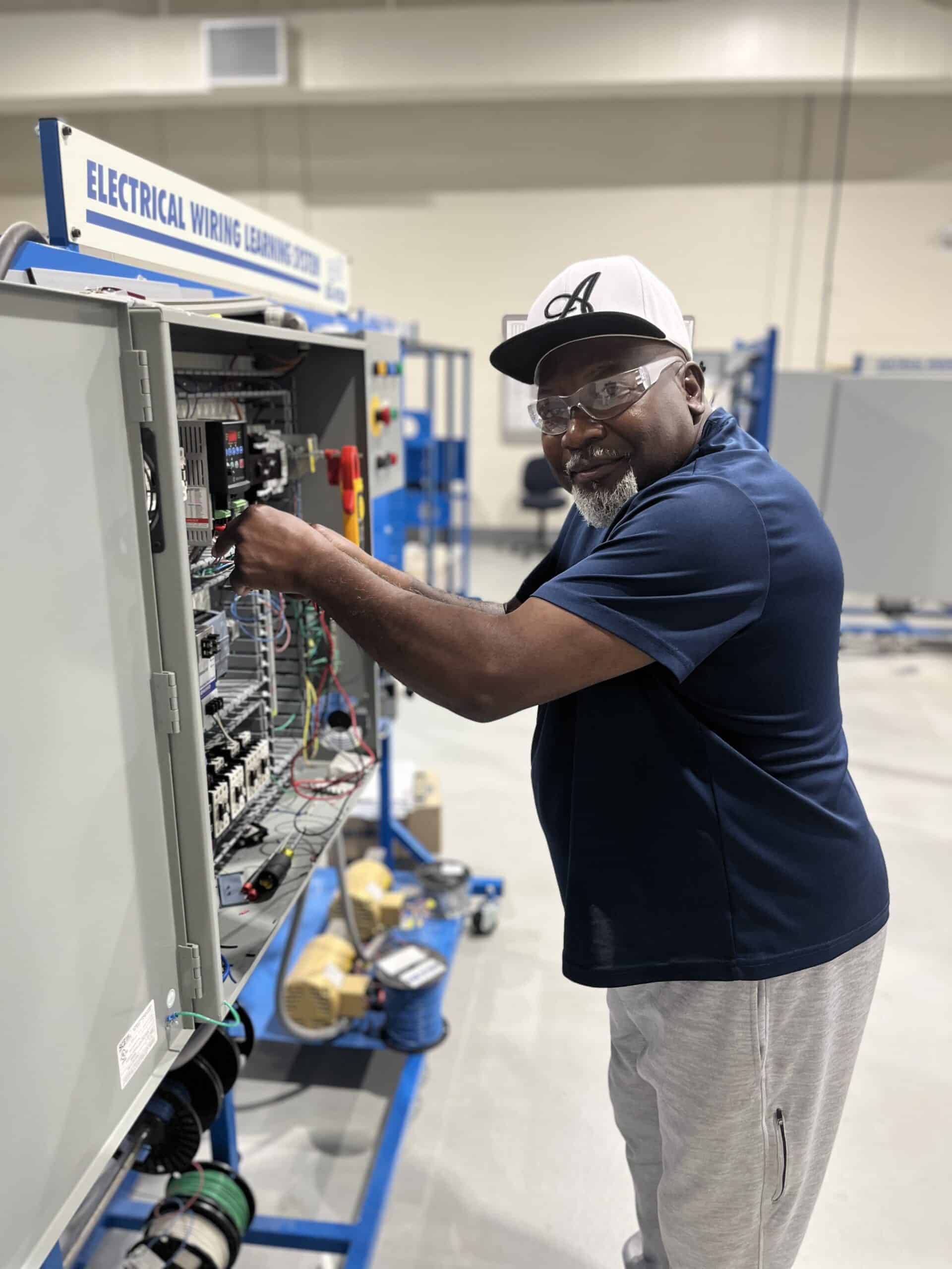 A man wearing glasses, a white and black cap, and a navy polo shirt works on an electrical wiring training panel in a workshop or classroom setting.
