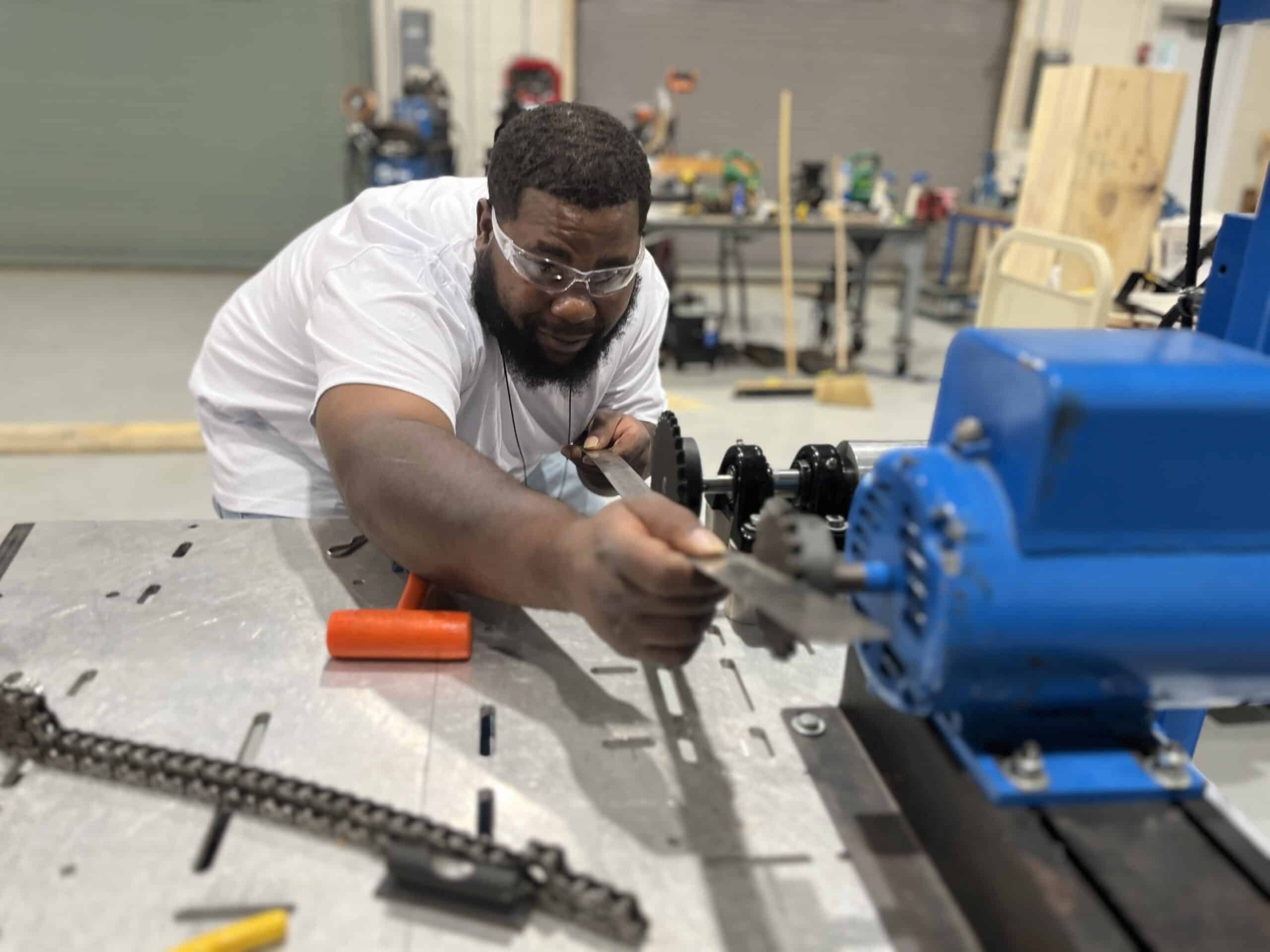 A man wearing safety goggles works with tools on a blue industrial machine in a workshop. Various tools and equipment are visible on the workbench and in the background.