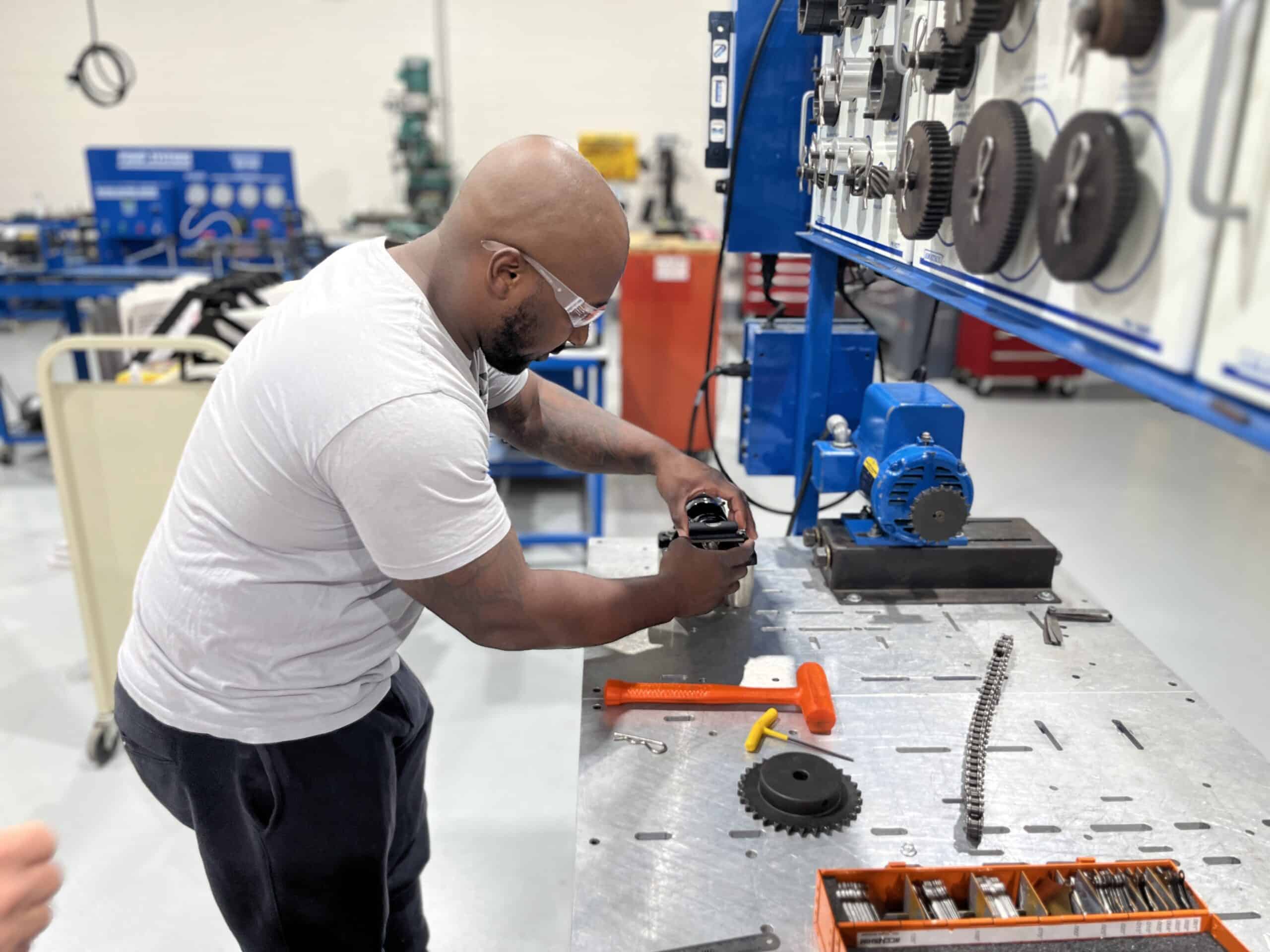 A man wearing safety glasses assembles mechanical parts at a workbench in a workshop, surrounded by gears, tools, and mechanical components.