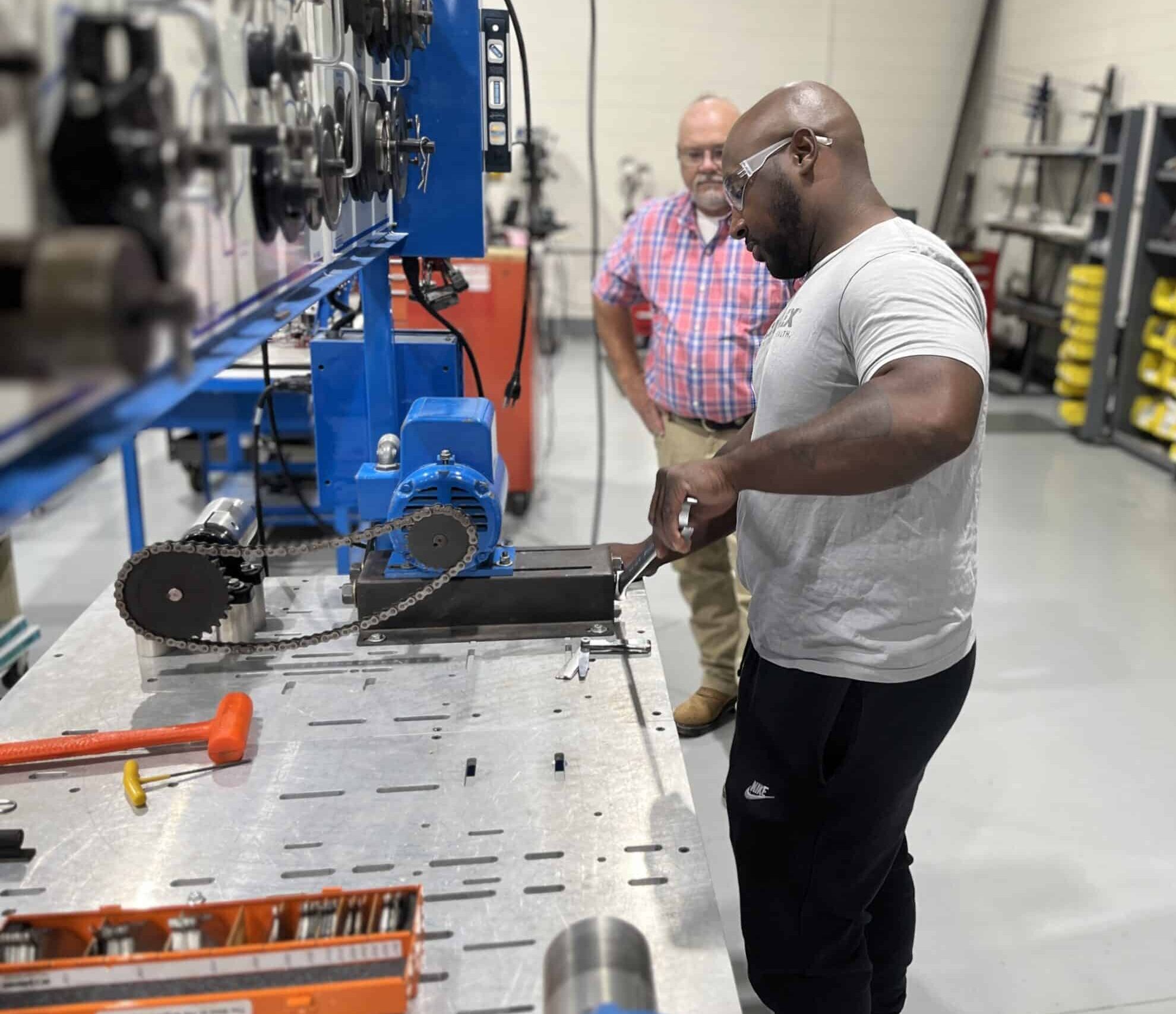 Two men are in a workshop; one is working with tools on a metal table, while the other observes. Various mechanical parts and equipment are visible in the background.