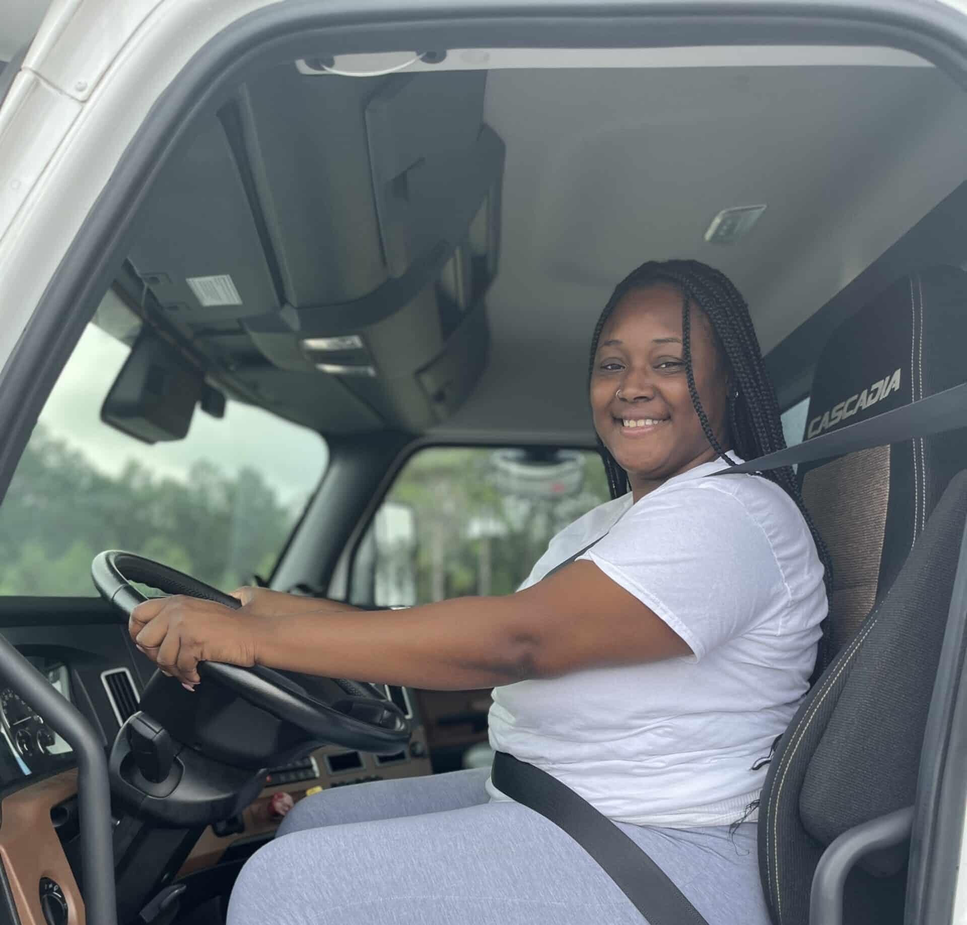 A smiling woman wearing a white t-shirt and gray pants sits in the driver’s seat of a large truck, holding the steering wheel and looking at the camera.