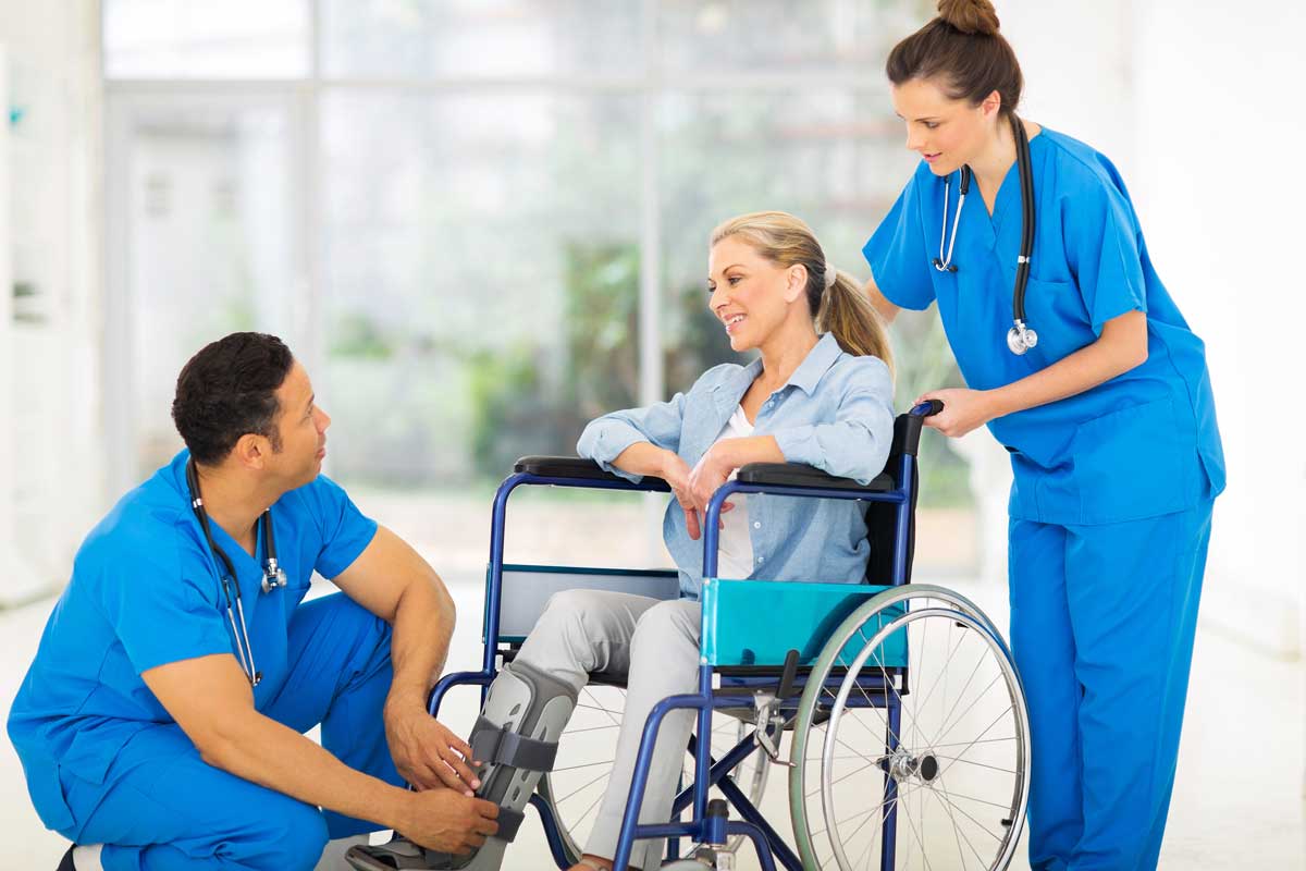 A woman in a wheelchair with a leg brace smiles while two healthcare professionals in blue scrubs attend to her, one crouching by her leg and the other standing and offering support.