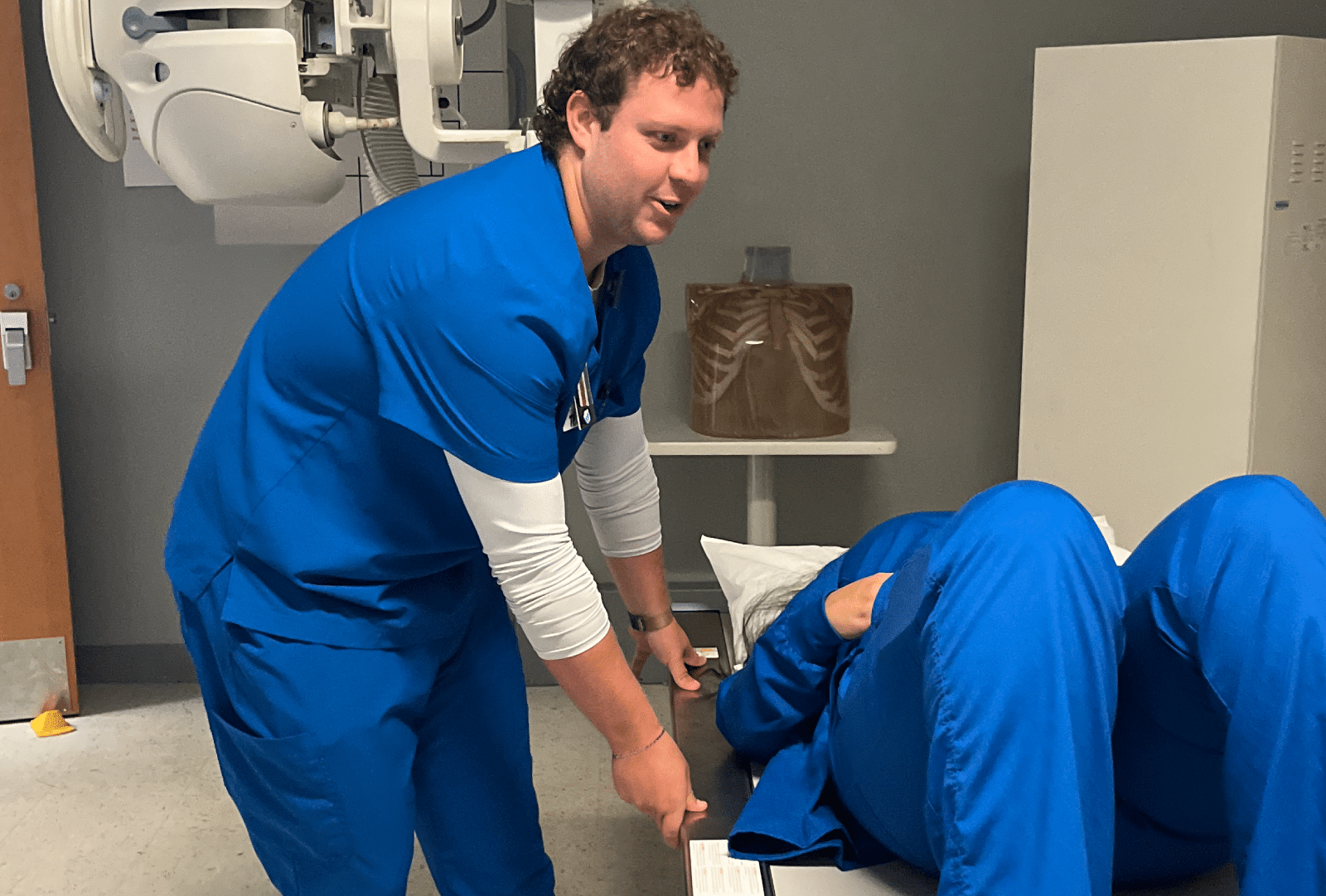 A healthcare worker wearing blue scrubs points at a clipboard as another person, also in blue scrubs, reclines on a medical bed with bent knees in a clinical examination room.