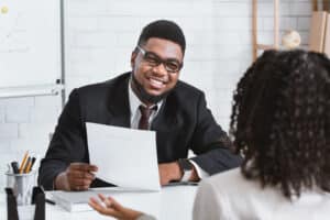 A man in a suit and glasses smiles while holding a resume and interviewing a woman across the table in a bright office setting.