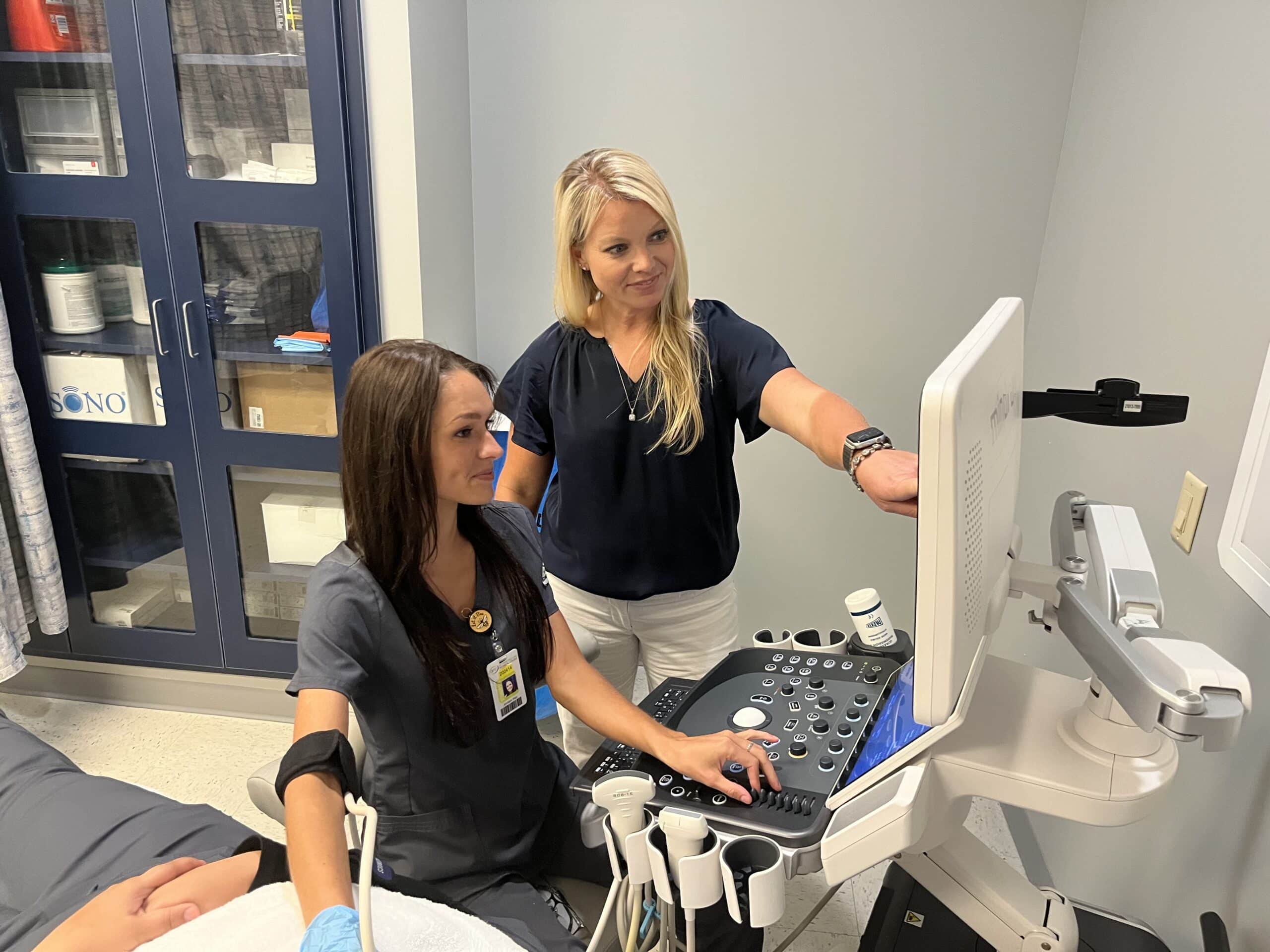 Two healthcare professionals operate an ultrasound machine in a medical examination room. One person is seated using the device on a patient while the other stands and points at the monitor, both focused on the screen.