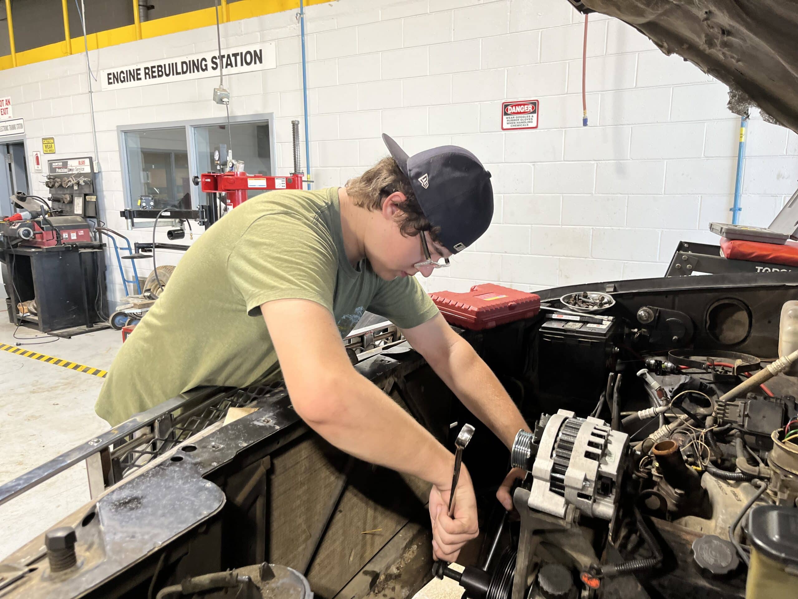 A person in a green shirt and backwards cap uses a wrench to work on a car engine in an automotive shop, with Engine Rebuilding Station sign visible in the background.