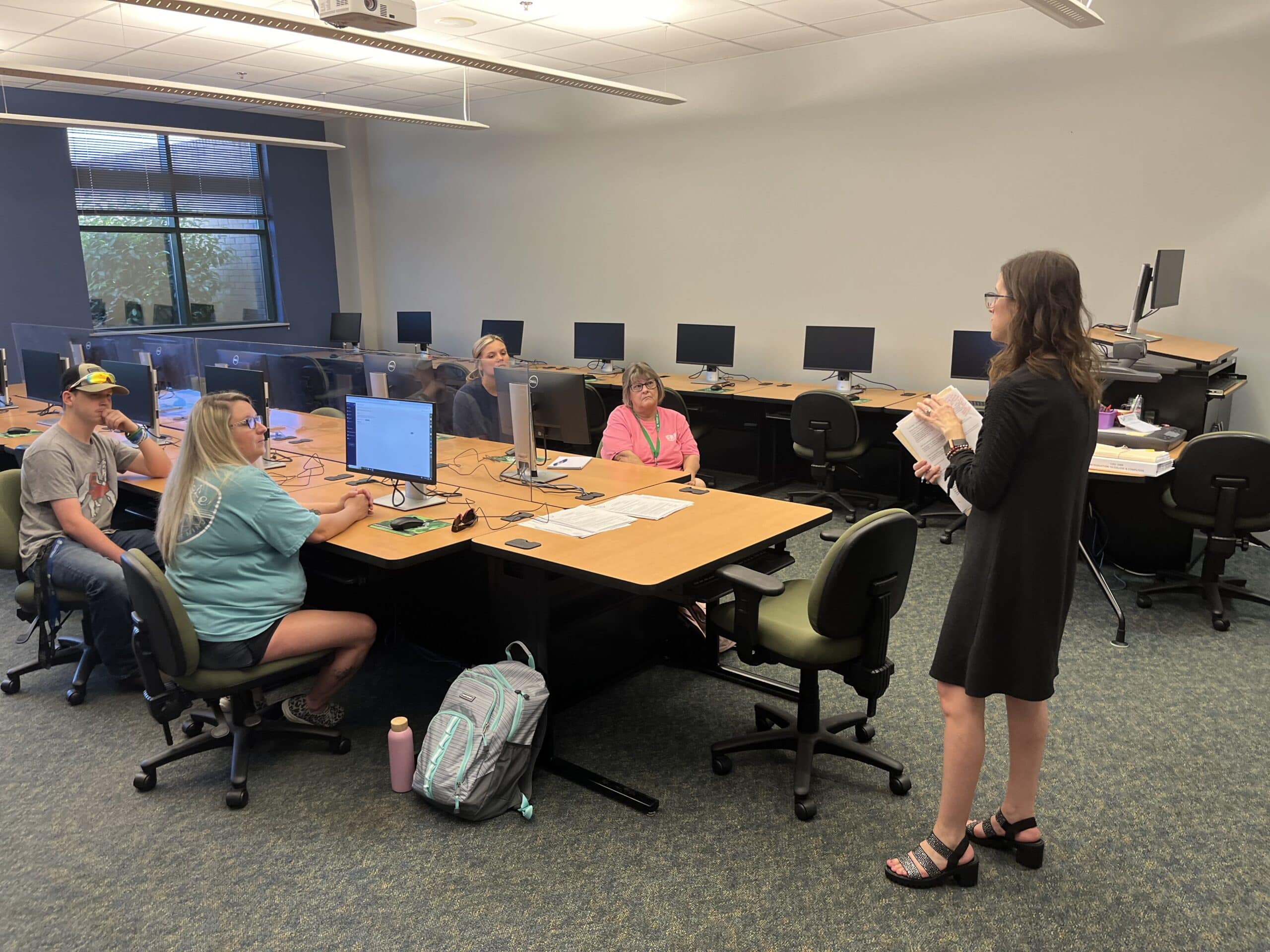 A woman stands and speaks to four seated adults in a computer lab classroom. The students listen attentively, with computers, papers, and backpacks on the desks. The room has large windows and overhead lights.