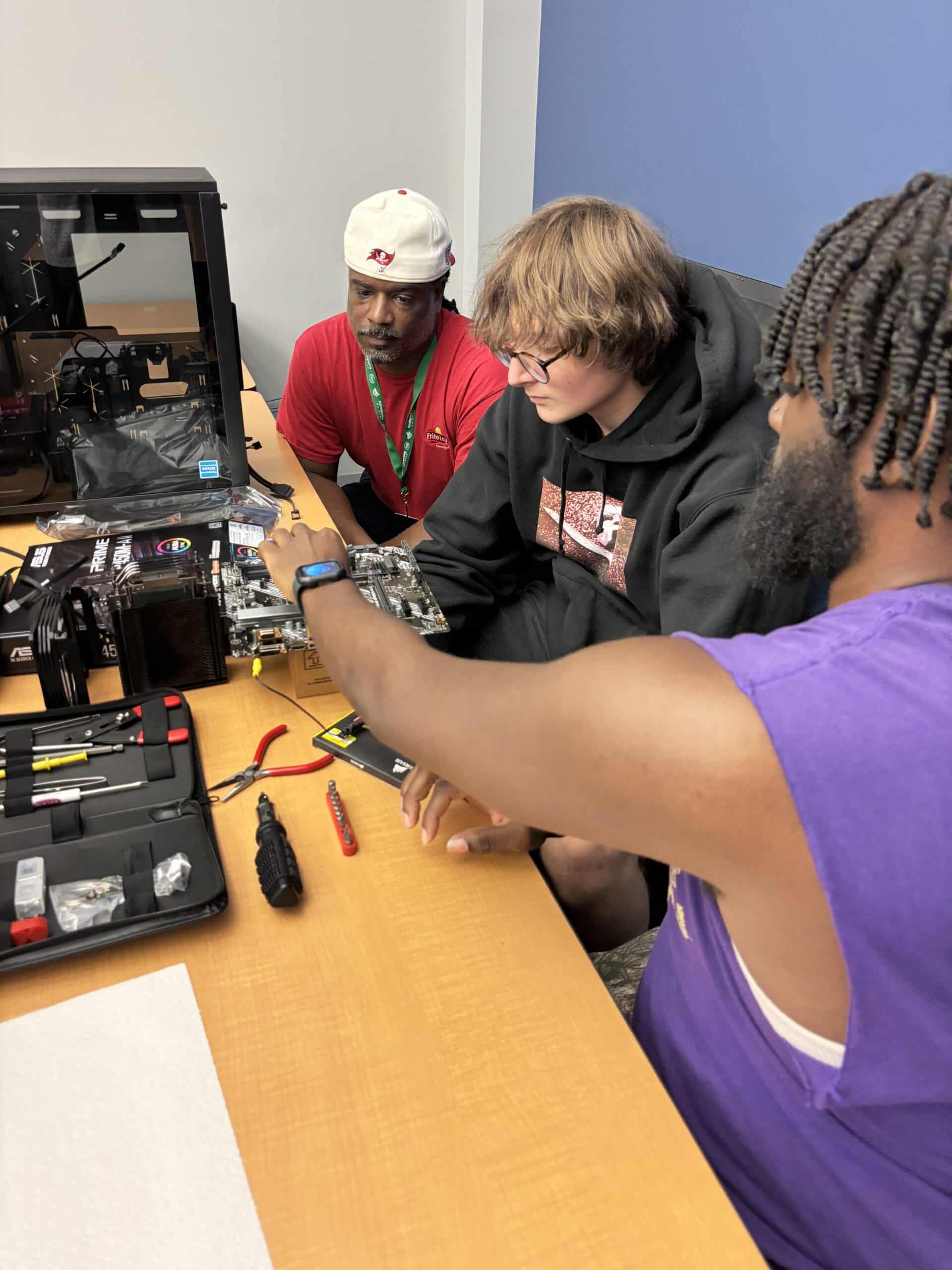 Three people sit around a table assembling computer hardware. One points inside a computer case while the others watch. Tools and components are scattered on the table, and a toolkit is open nearby.