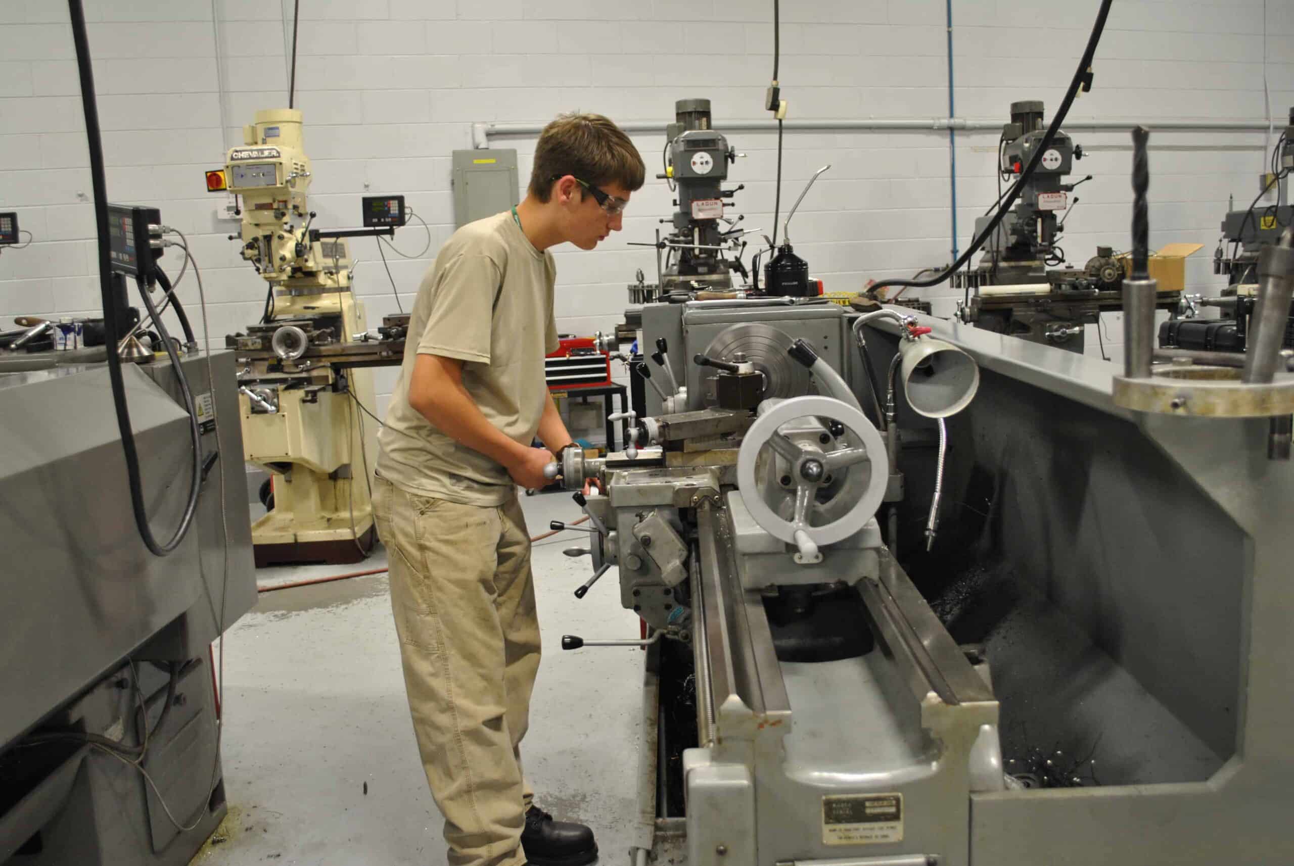 A person wearing safety glasses and beige clothing operates a metal lathe in a machine shop filled with industrial equipment and milling machines.