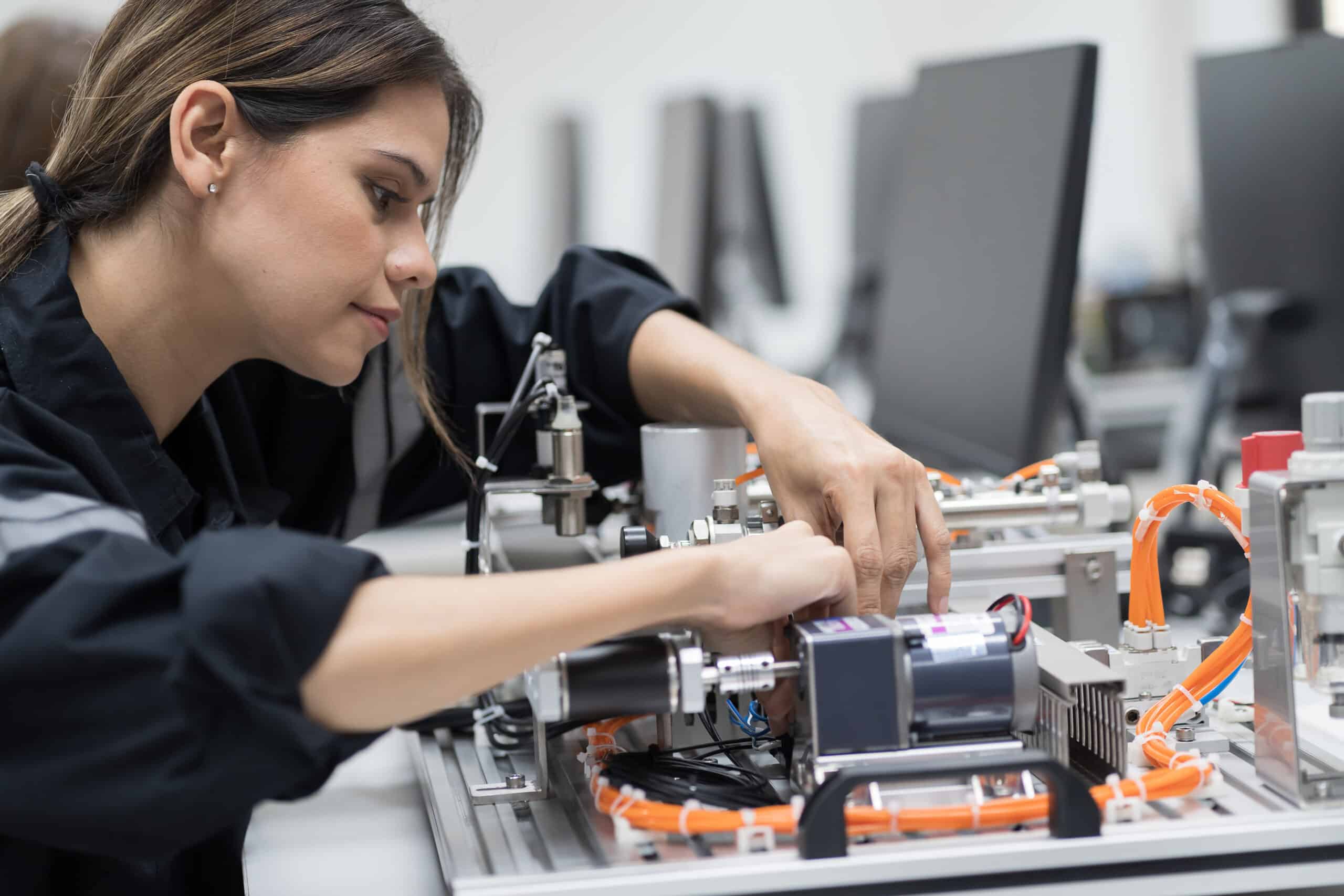 A woman wearing a dark jacket works intently on assembling or adjusting components of an electronic or mechanical device in a lab with computers and equipment in the background.