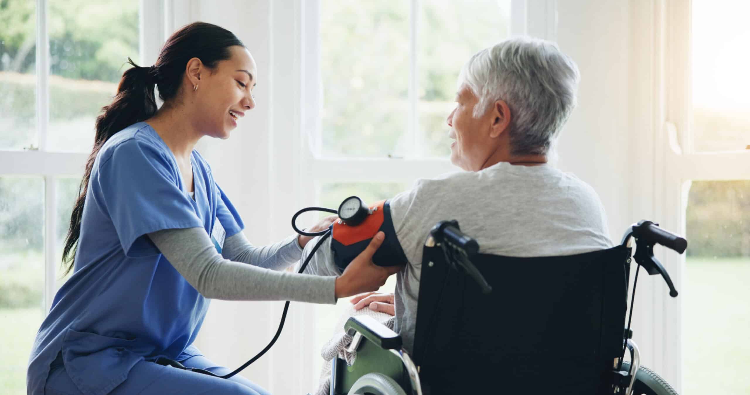 A nurse in blue scrubs smiles while taking the blood pressure of an older adult in a wheelchair by large, sunlit windows.