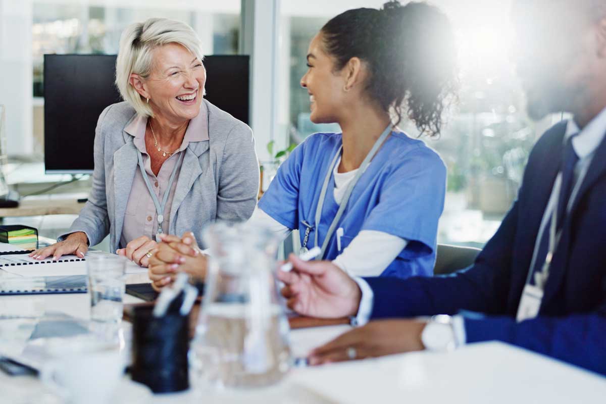 Two women, one in business attire and one in scrubs, smile and talk at a meeting table with a man in a suit. Papers, water glasses, and office supplies are on the table in a bright, modern office.
